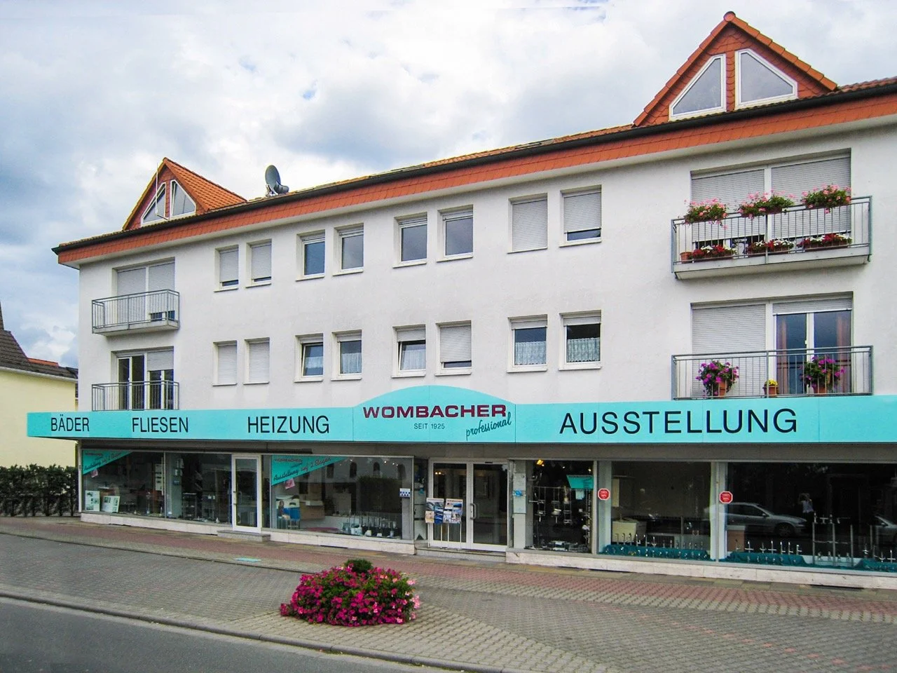 Ein Mehrfamilienhaus mit Geschäften im Erdgeschoss, die eine Ausstellung für Badezimmer, Fliesen und Heizungen anbieten, mit Fensterblumen und einem Blumenbeet vor der Straße.