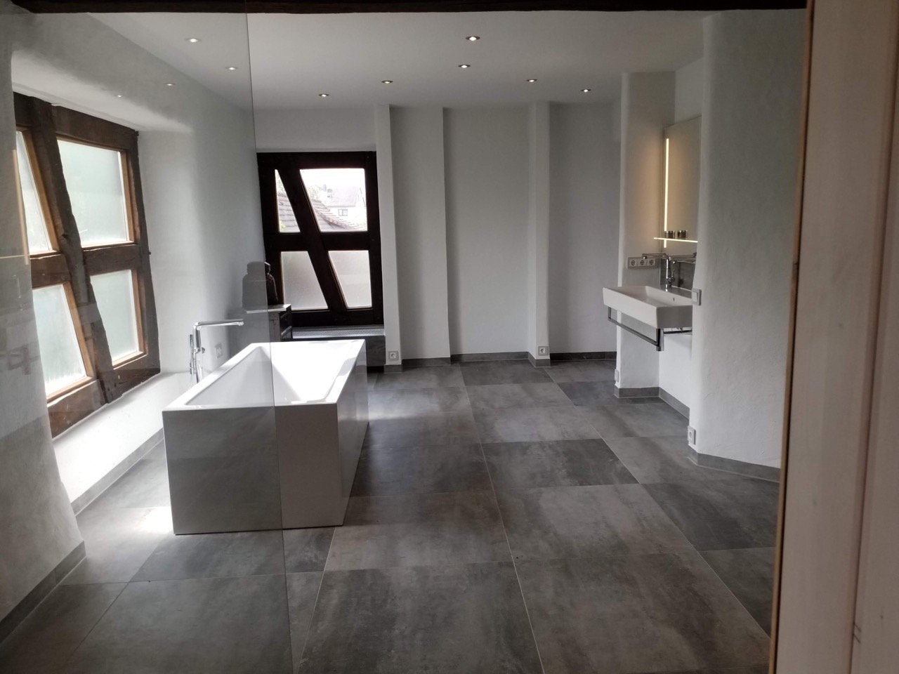 Modernes Badezimmer mit Badewanne, Waschbecken, großen Fenstern und Holztüren, hell und minimalistisch gestaltet.