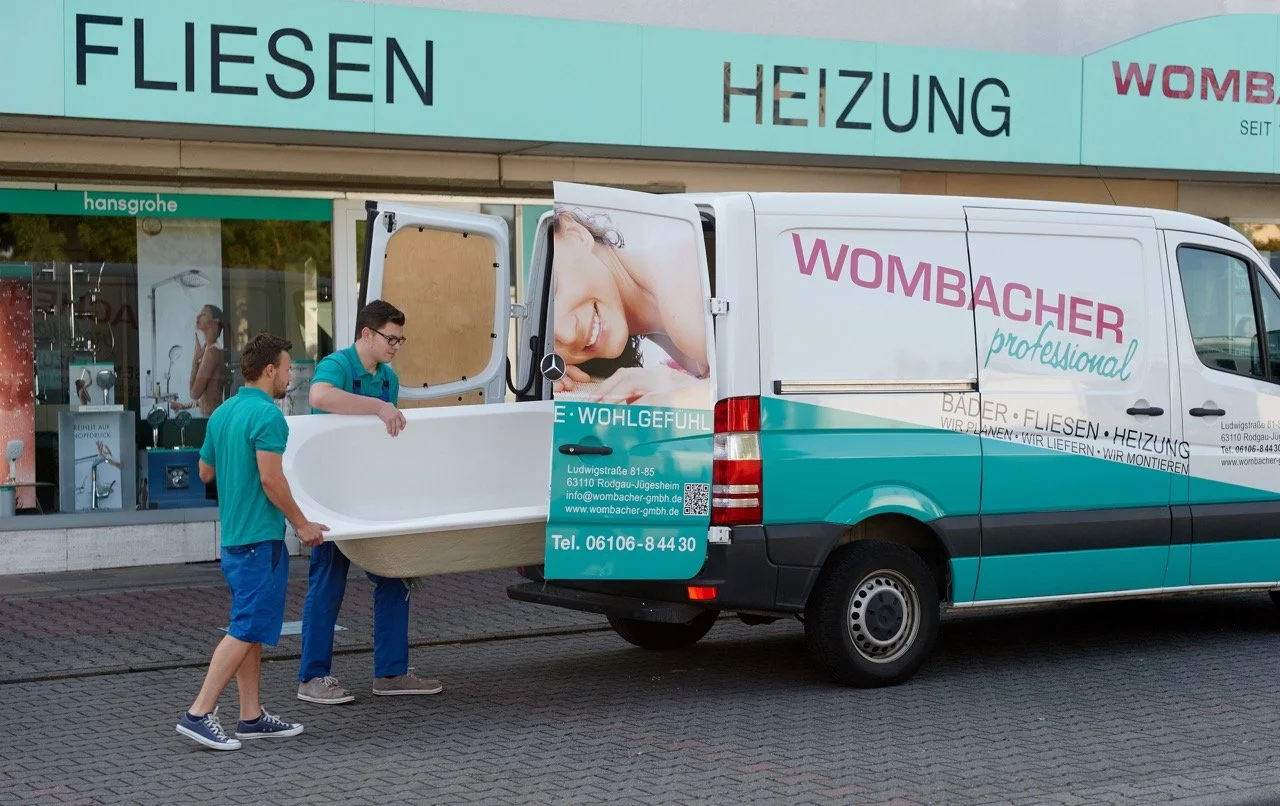 Zwei Männer in blauen Uniformen tragen eine Badewanne in einen Lieferwagen mit der Aufschrift 'Wombacher professional'. Im Hintergrund ein Ladengeschäft mit Schildern für Fliesen und Heizung.