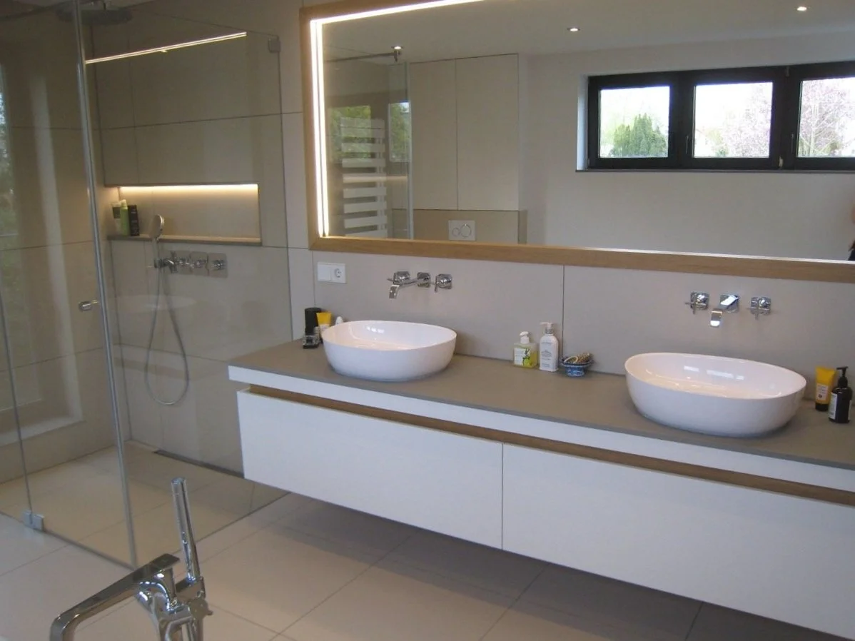 Moderne Badezimmer mit Doppelwaschbecken, Spiegel mit Beleuchtung, Duschbereich mit Glaswand, Fenster und weißen Fliesen.