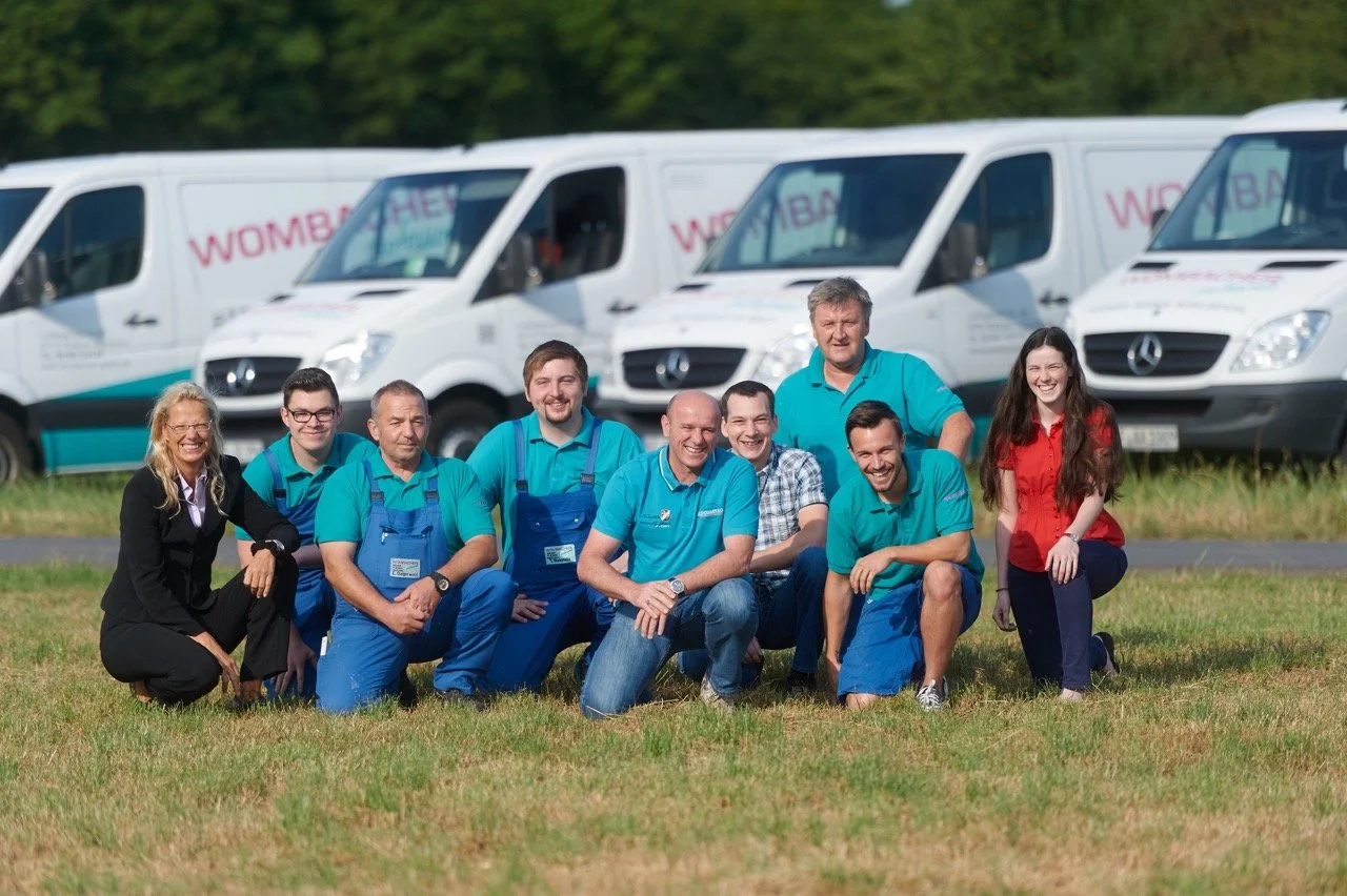 Gruppe von Menschen in blauer Kleidung, die vor weißen Lieferwagen im Freien posieren.