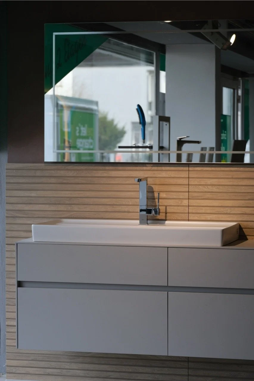 Ein weißes Waschbecken auf einem grauen Waschtisch vor einer Wand mit Holzvertäfelung, darüber befindet sich ein senkrechter Spiegel mit einem Blick auf die Fenster und eine Dachterrasse im Hintergrund.