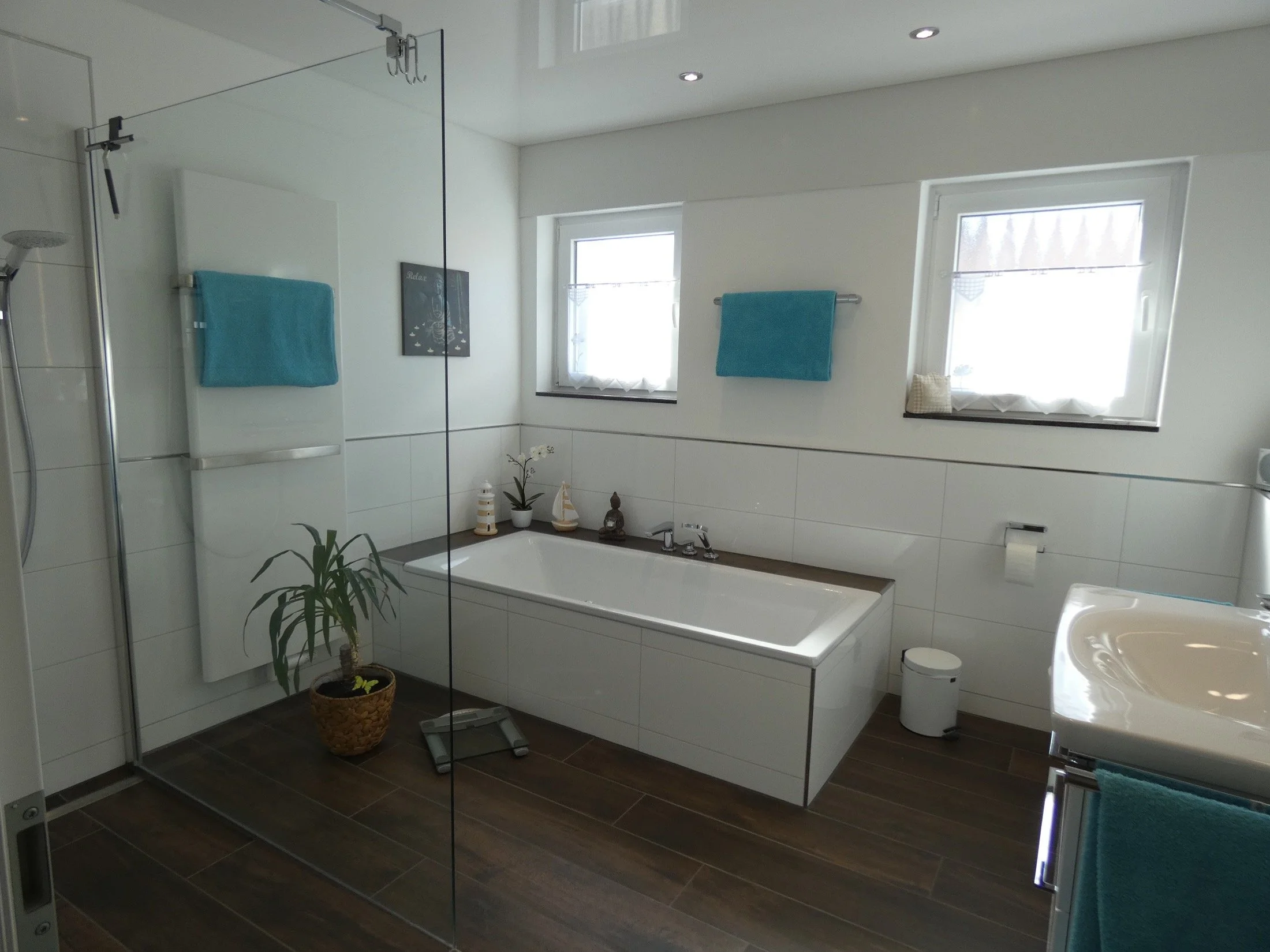 Geräumiges Badezimmer mit Badewanne, Dusche, zwei Fenster, blaue Handtücher, Pflanze und Deko-Elemente.