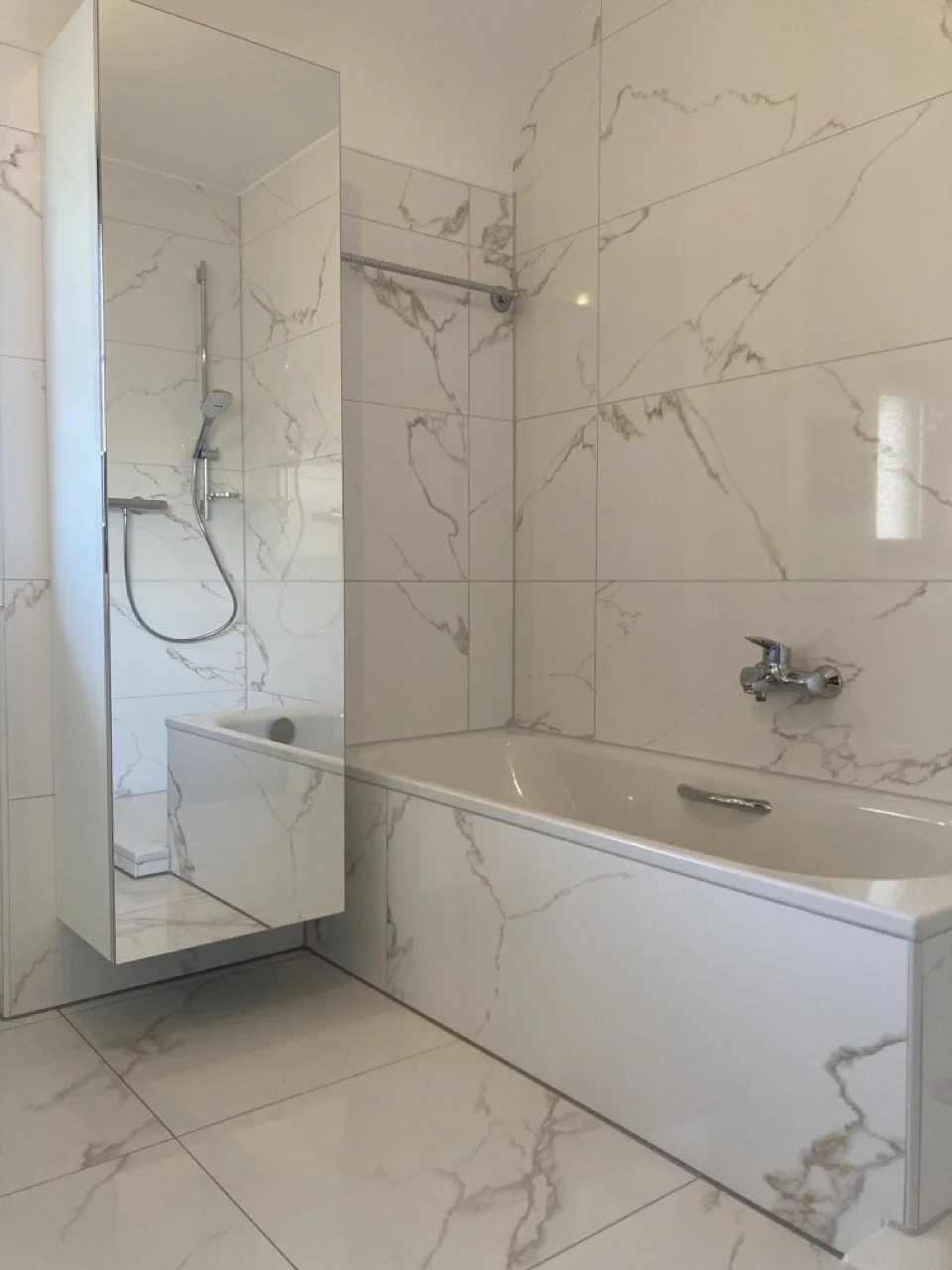 Badezimmer mit weißen Marmorfliesen, Badewanne, Wasserhahn, Dusche mit Handbrause, Spiegel