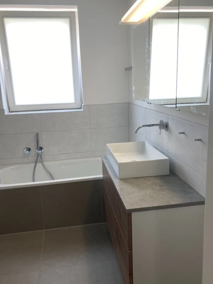 Ein Badezimmer mit einem weißen Waschbecken auf einer grauen Arbeitsplatte, einem Spiegel mit Schrank, einer Badewanne, zwei Fenster und neutralen Wandfliesen.