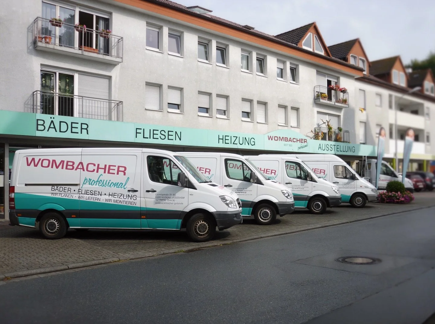 Mehrere weiße Firmenwagen mit WOMBACHER Logo, die auf einem Parkplatz vor einem Geschäft mit Schaufenster stehen, das Fliesen, Bäder, Heizung und Ausstellung in Schildern anzeigt.