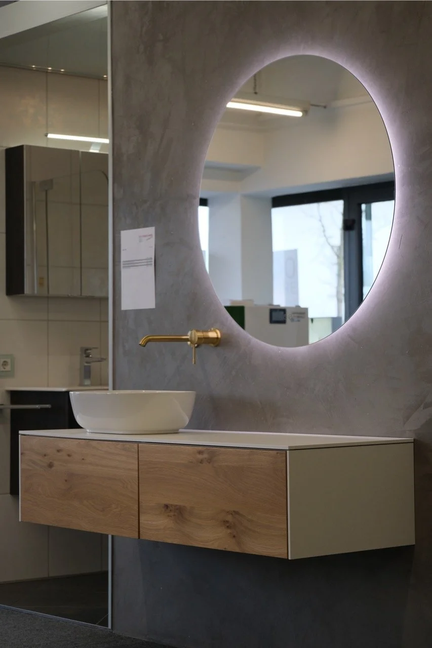 Moderne Badezimmerwand mit großem runden Spiegel, einem weißen Waschbecken auf einem Holztisch, goldener Wasserhahn, Hintergrund mit Fenstern und beleuchteter Wand.