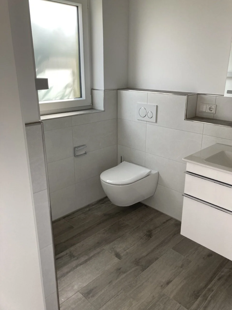 Modernes Badezimmer mit weißem wandhängendem WC, grauen Fliesen, einem Fenster und einer weißen Waschtisch mit Schubladen.