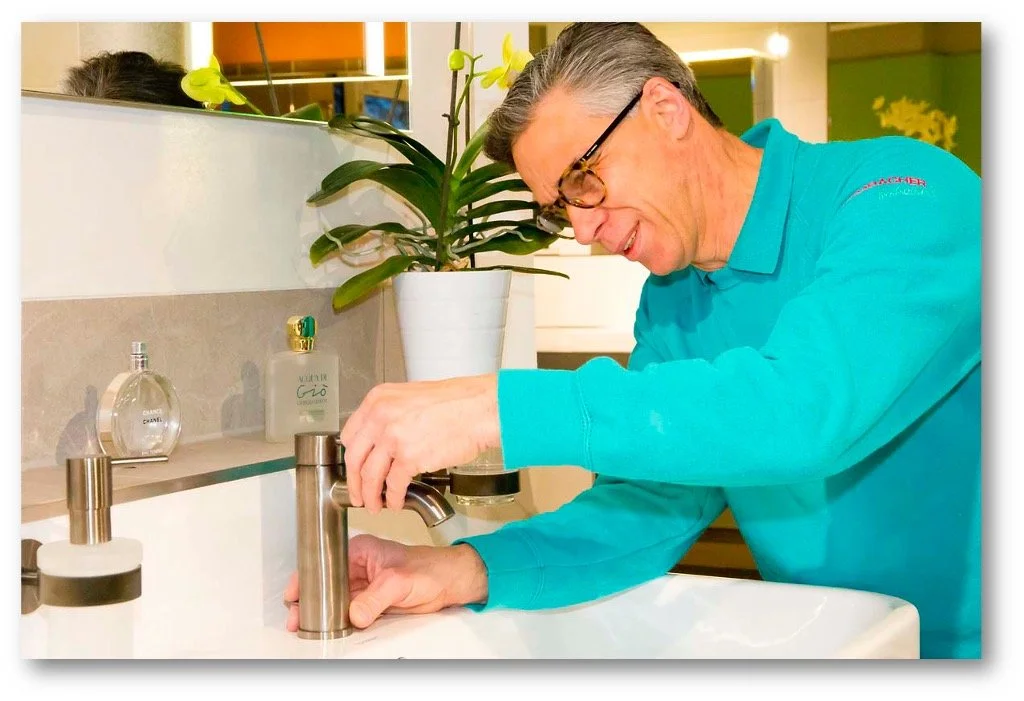 Ein Mann mit Brille in einem blauen Pullover arbeitet an einer Wasserhahn in einem Badezimmer, neben einer Orchidee in einer weißen Topfpflanze.