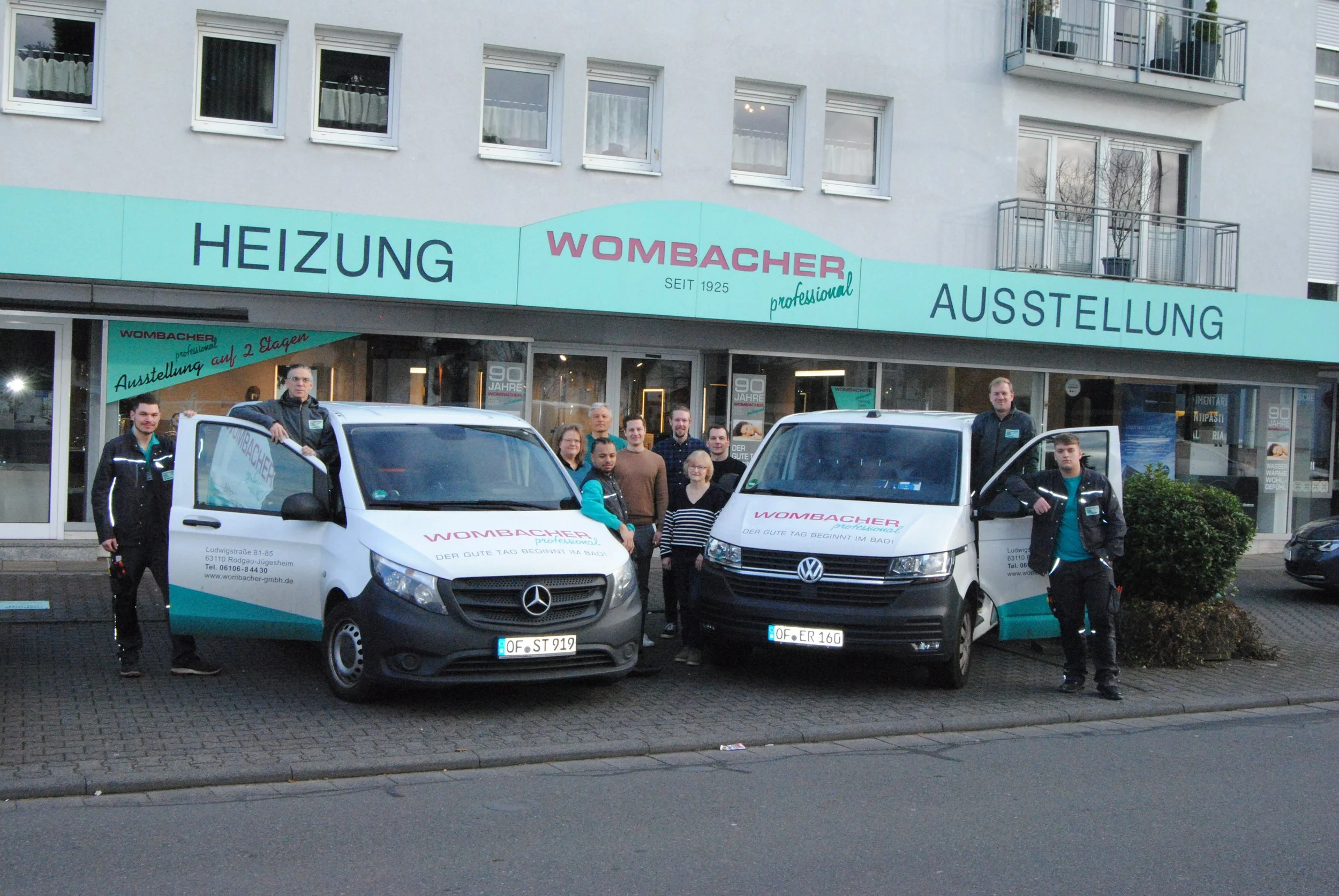 Eine Gruppe von Menschen steht vor zwei Firmenfahrzeugen vor einem Geschäft mit der Aufschrift "Wombacher" und "Heizung Ausstellung". Die Fahrzeuge sind weiße Lieferwagen mit der Markenbeschriftung." Wombacher" und "der gute Tag beginnt im Bau".