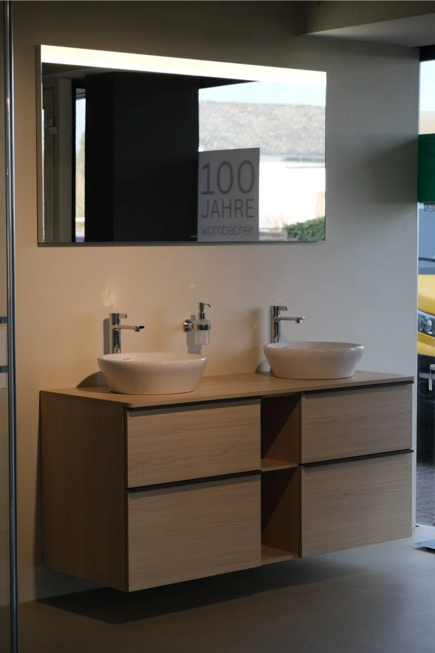 Doppelwaschbecken mit zwei Wasserhähnen auf einer Holzkonsole, großer Wandspiegel darüber, im Hintergrund ein Fenster mit Blick nach draußen, auf dem Spiegel befindet sich ein Schild mit der Aufschrift "100 Jahre Wombacher".