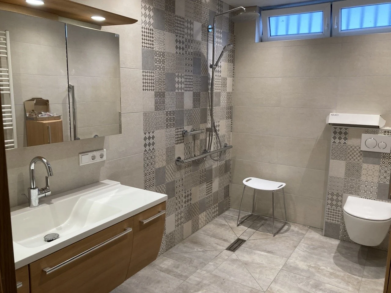 Badezimmer mit Waschbecken, Spiegel, Dusche und WC, beige Fliesen, Fenster an der Decke, kleiner Stuhl, Heizkörper