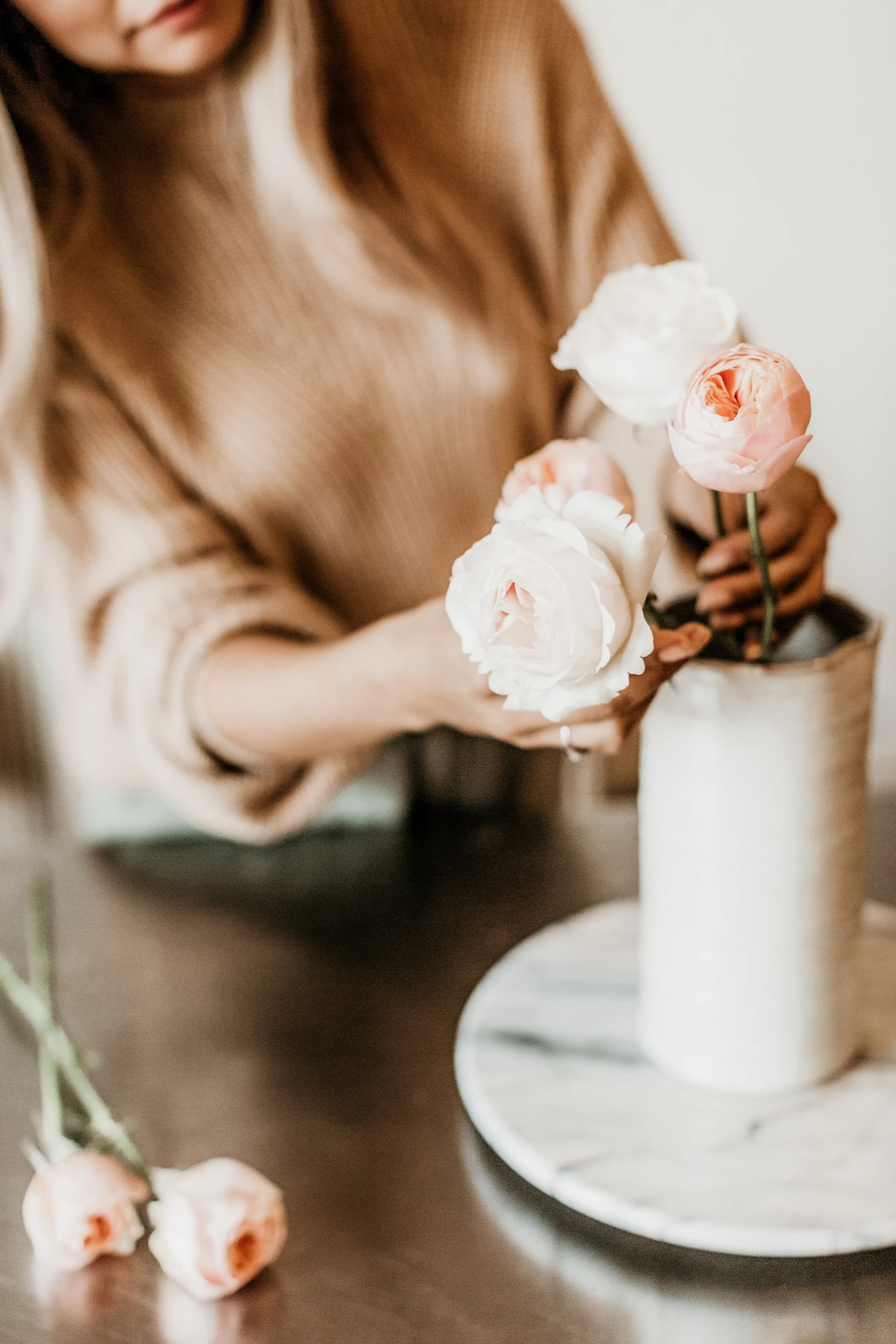 A woman arranging white and pink peonies in a ceramic vase on a table.