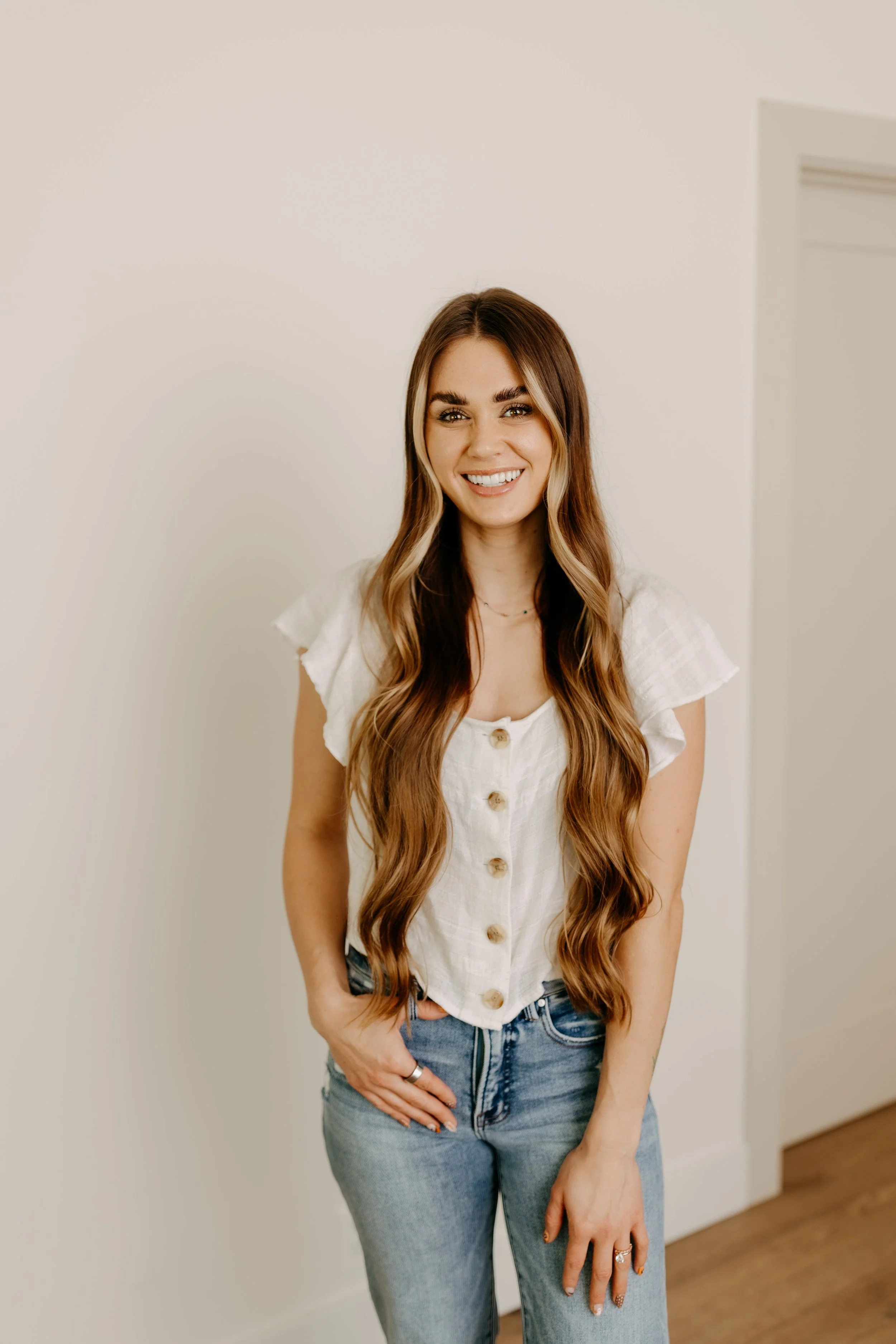 A young woman with long wavy brown hair wearing a white button-up top and blue jeans, smiling and standing indoors against a plain white wall.