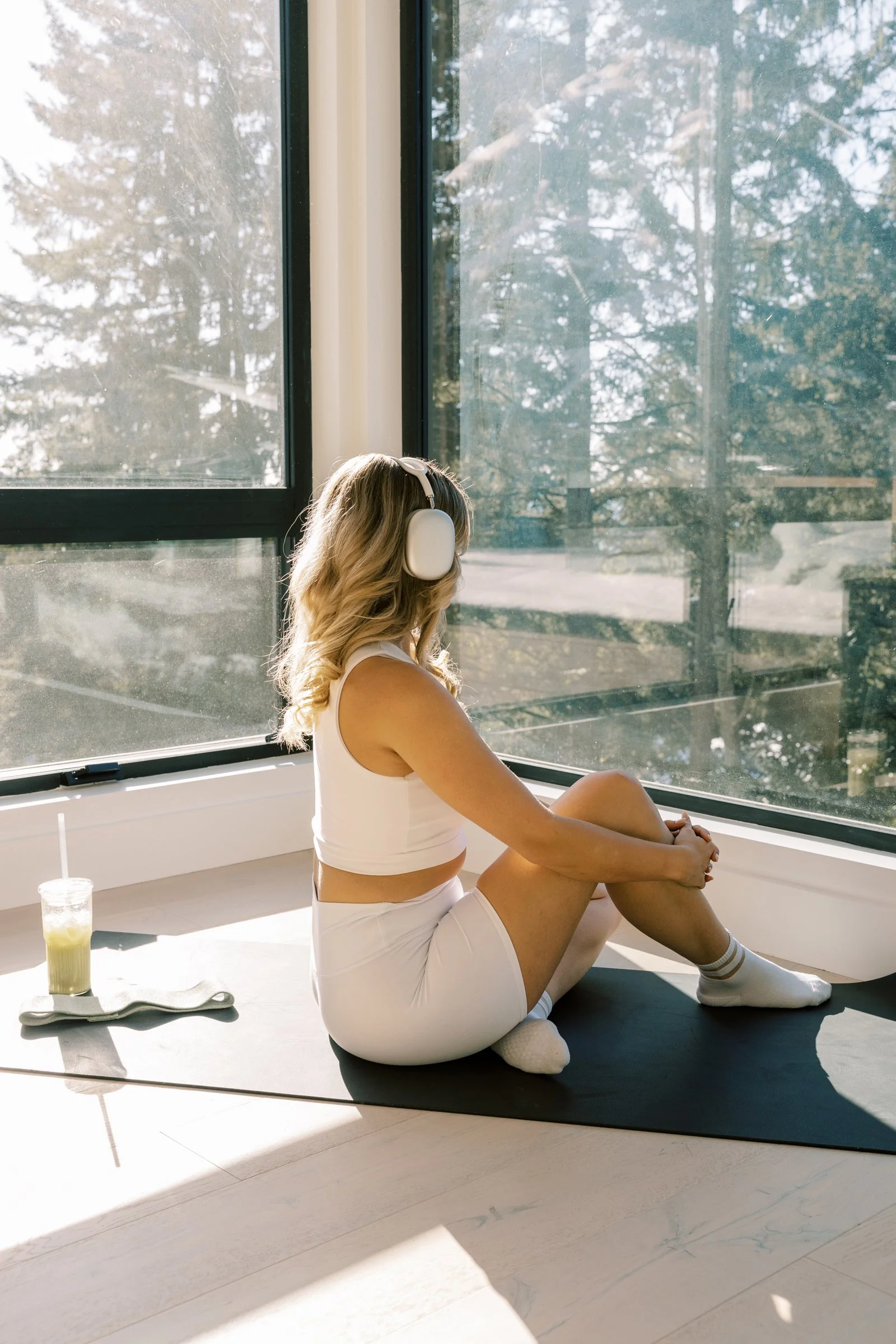 A woman sitting on a yoga mat, wearing workout clothes and headphones, next to a large window with sunlight streaming in.