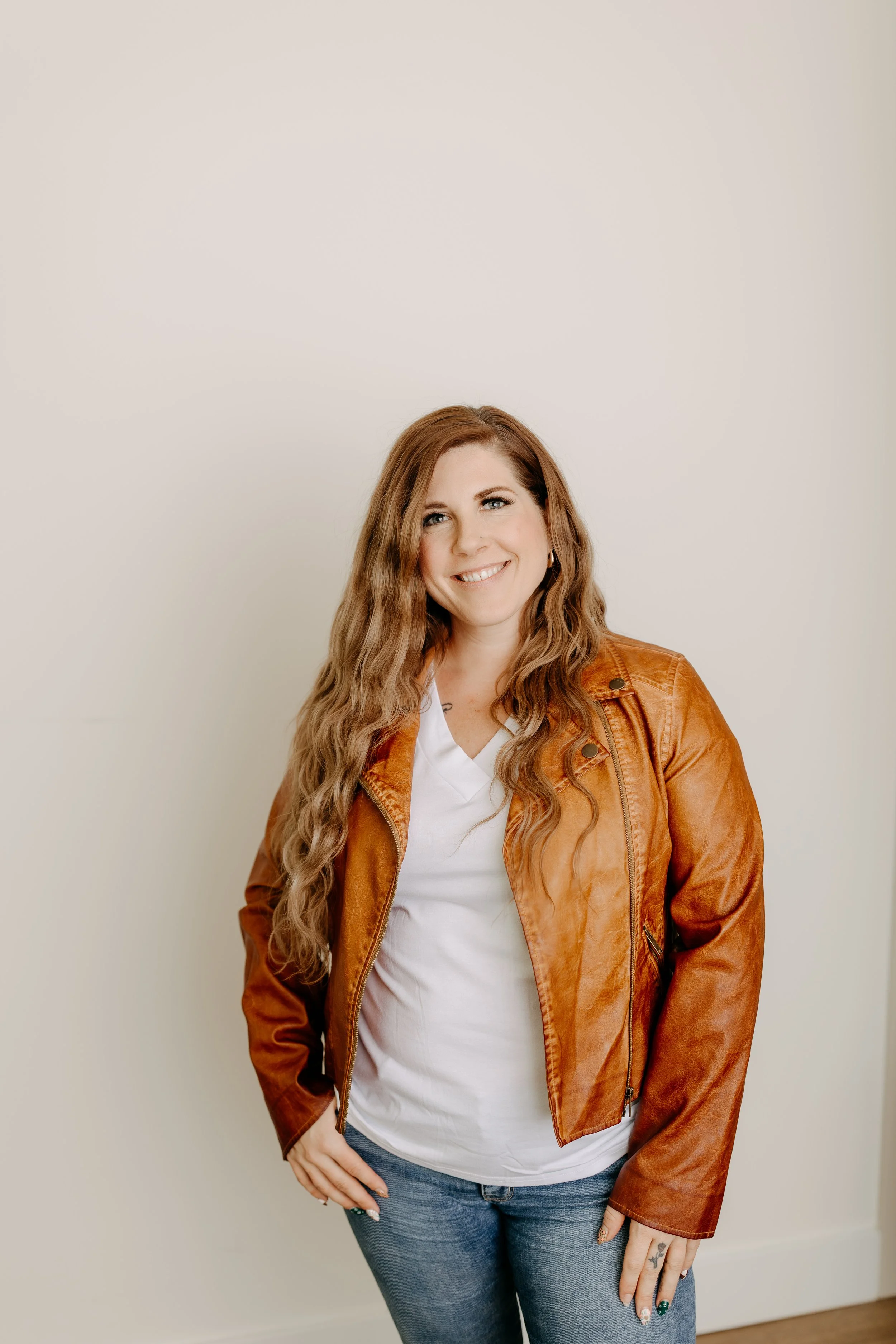 A woman with long wavy auburn hair smiling and standing with hands on her hips, wearing a brown leather jacket, white t-shirt, and blue jeans against a plain light-colored wall.