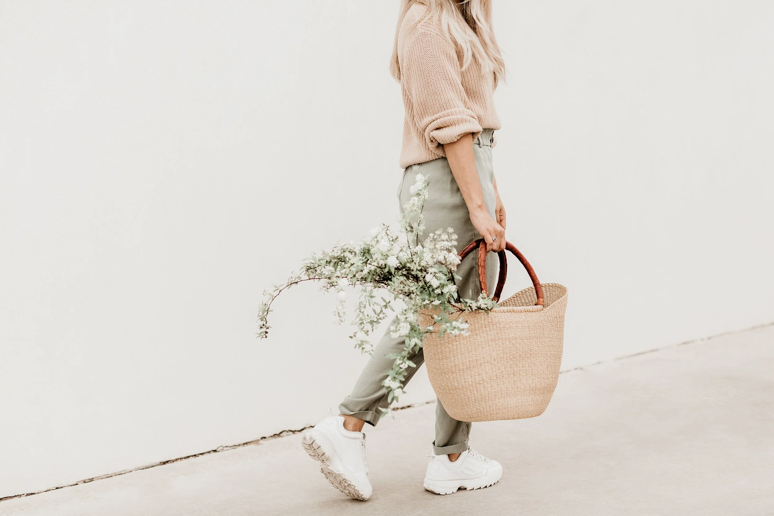 Person dressed in beige sweater, green pants, and white sneakers walking with a large woven beige tote bag filled with white flowers, against a plain white wall