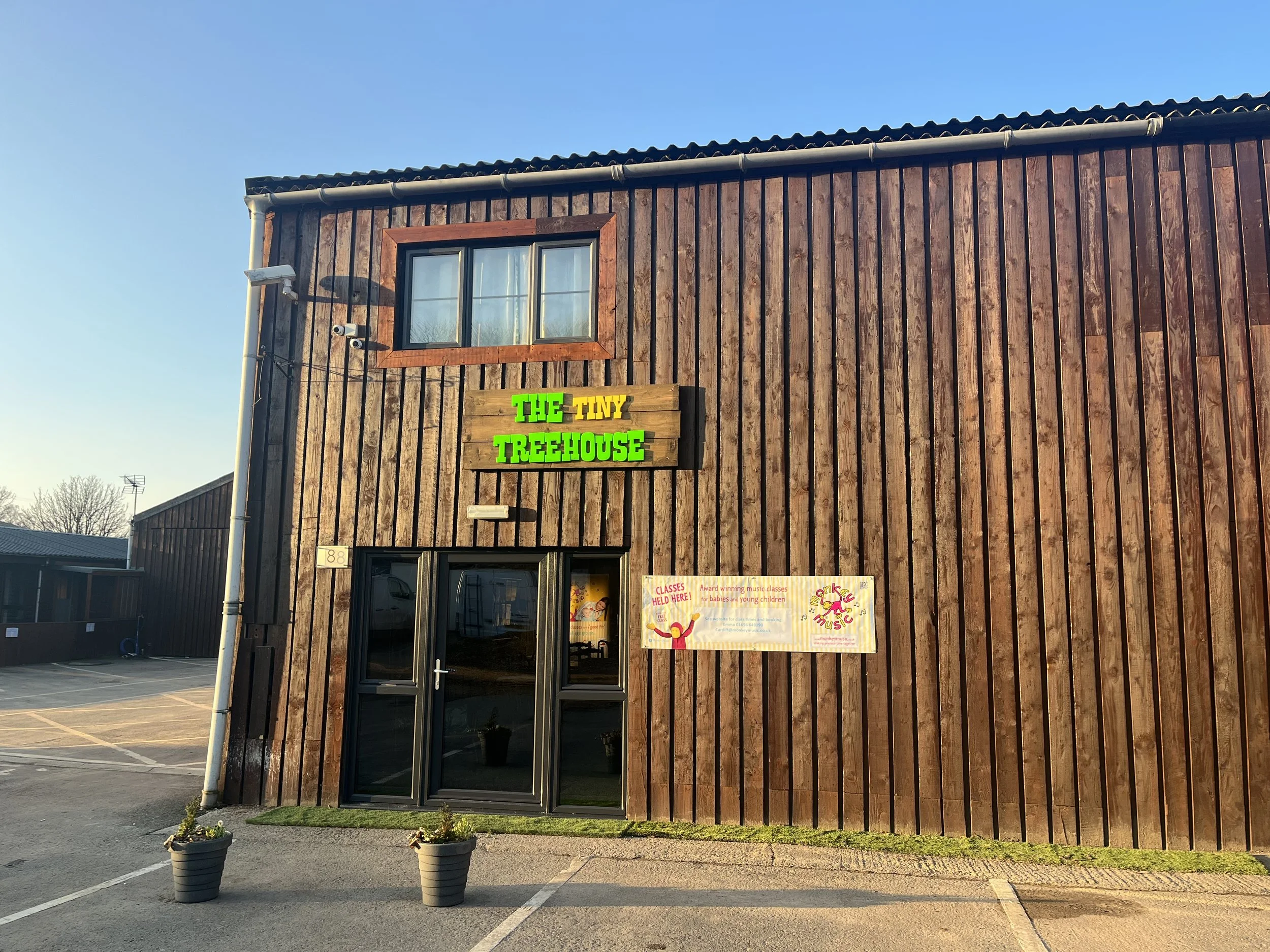 Wooden building labeled 'The Tiny Treehouse' with a sign advertising classes, and two potted plants near the entrance.