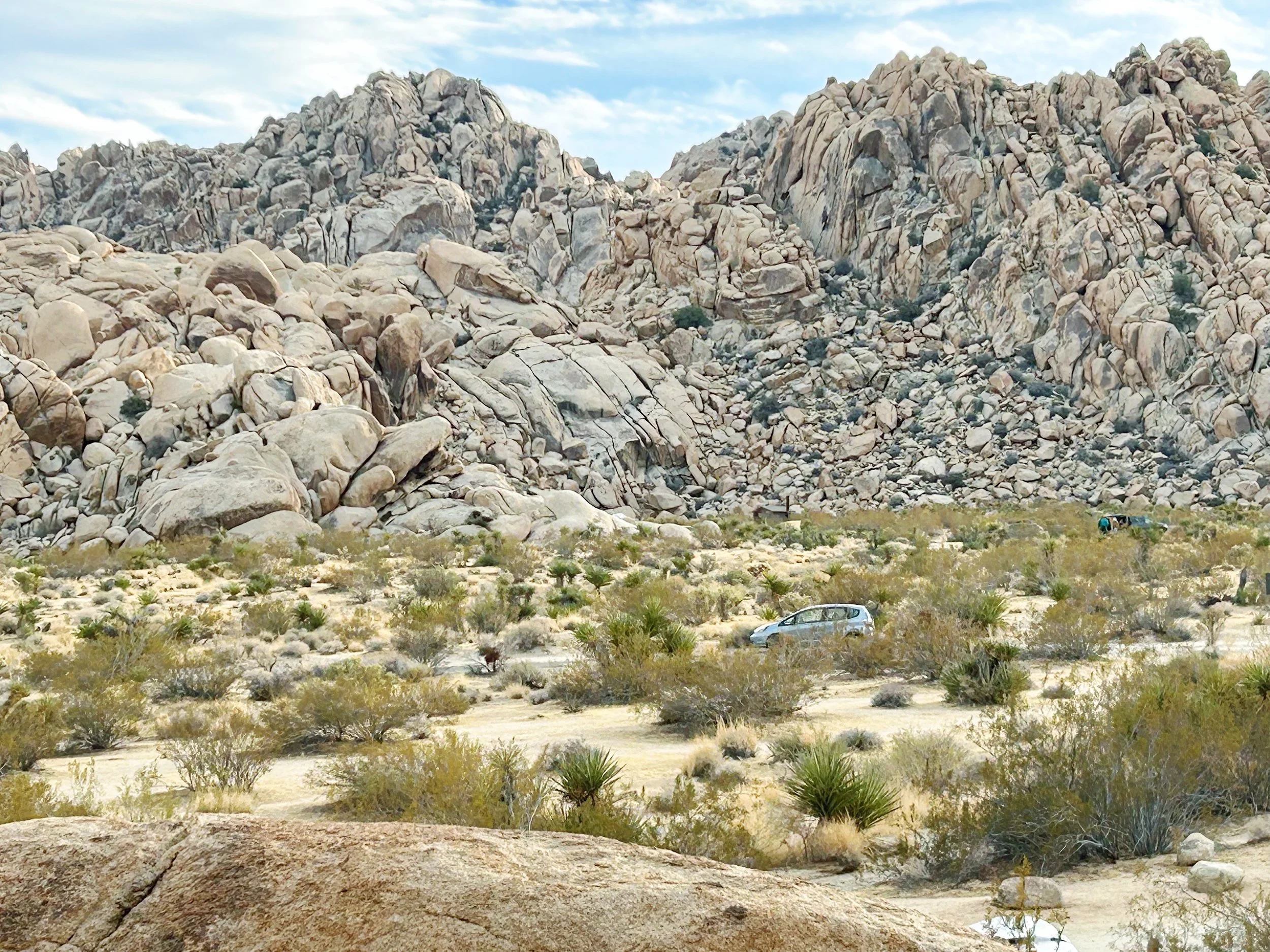 Indian Creek Camp Ground, in Joshua Tree National park. Desert landscape with large rocky mountains, sparse shrubs, and a few cars parked on the sandy terrain.