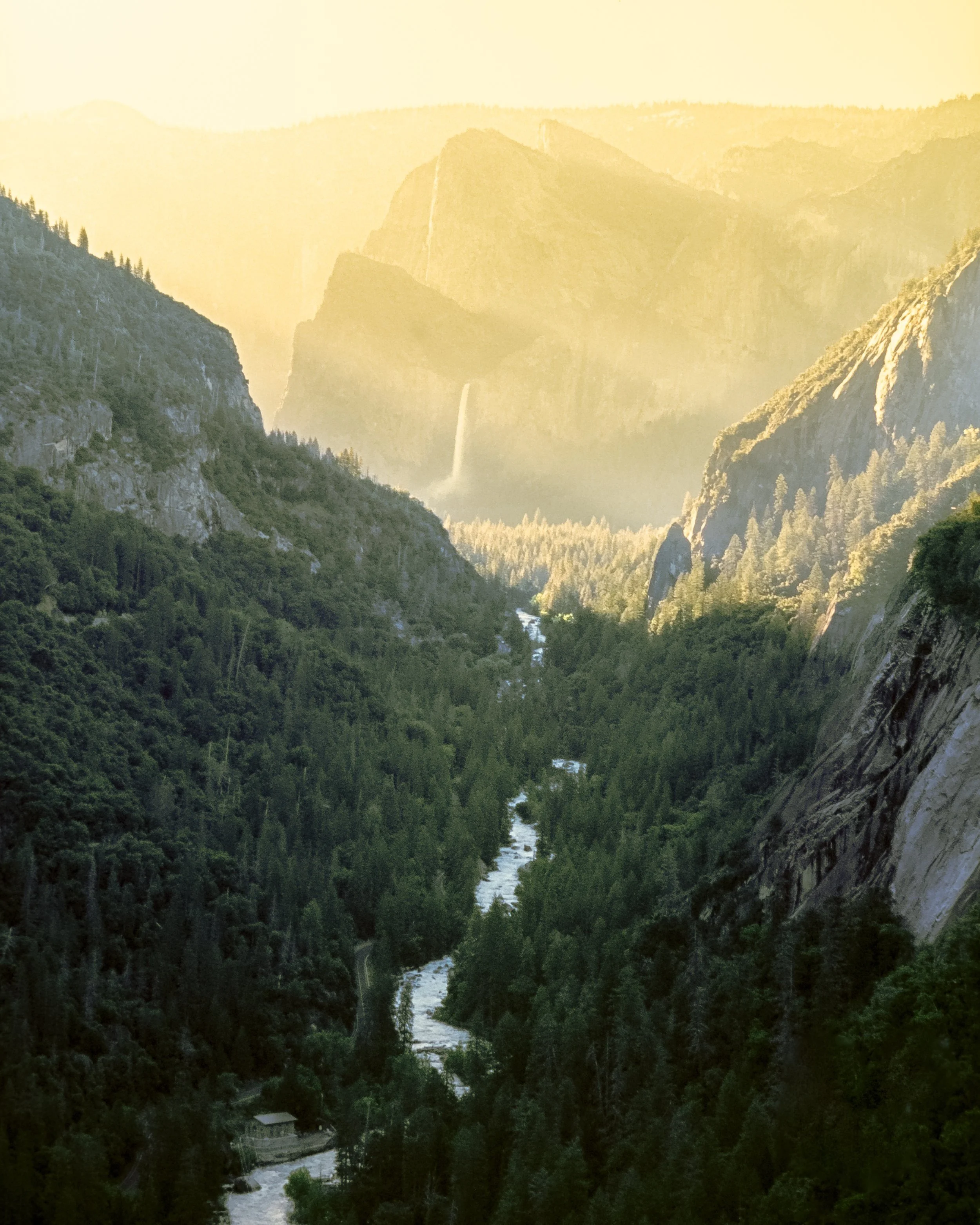 View from Separate Reality climb in Yosemite National Park — a sunlit lower Merced Canyon winding through, lush forest and misty waterfalls in the distance.