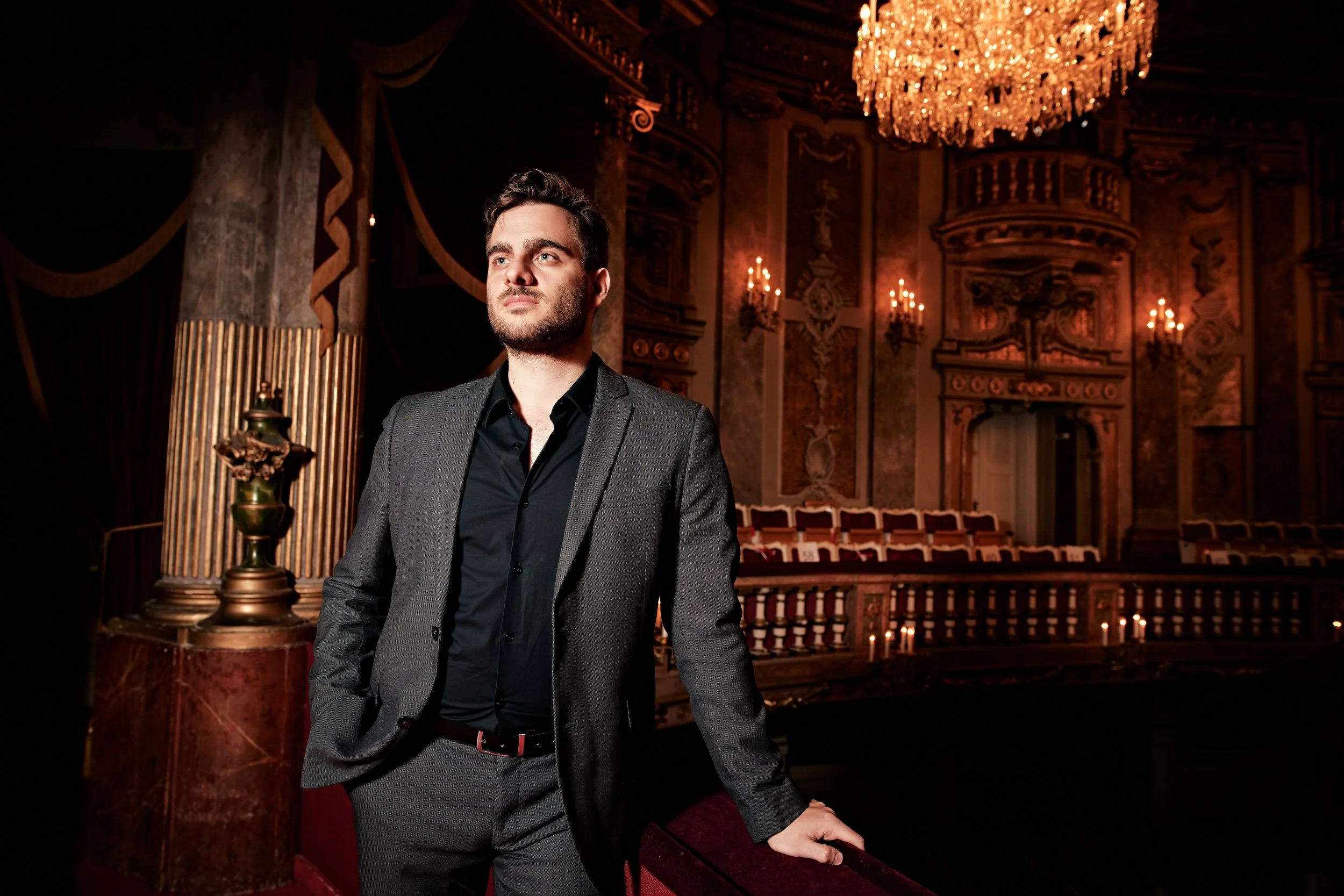 A man in a dark suit standing in an ornate theater with a large chandelier and gold accents.