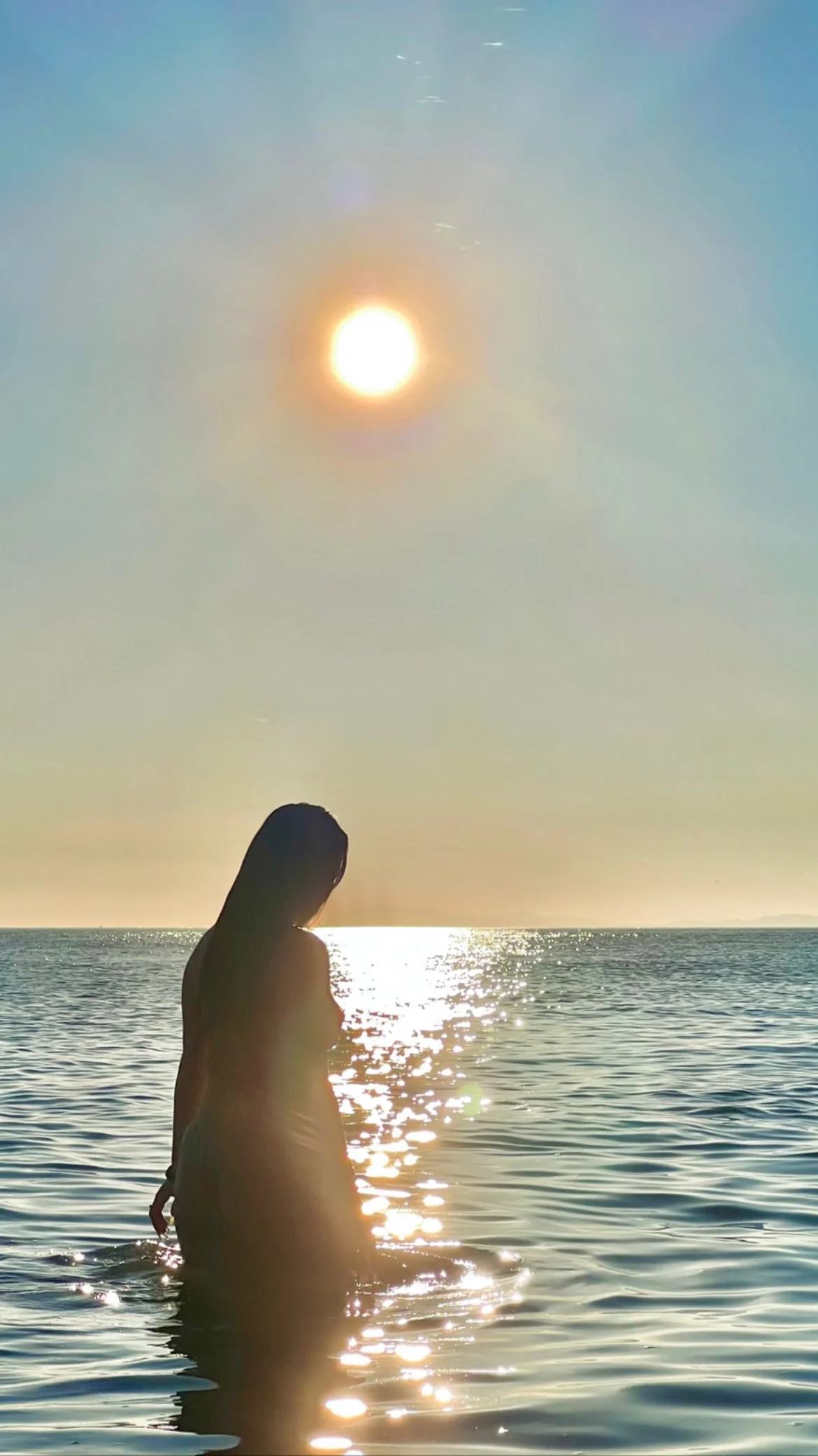 Silhouette of a woman standing in water at sunset, with the sun low in the sky and reflecting on the water