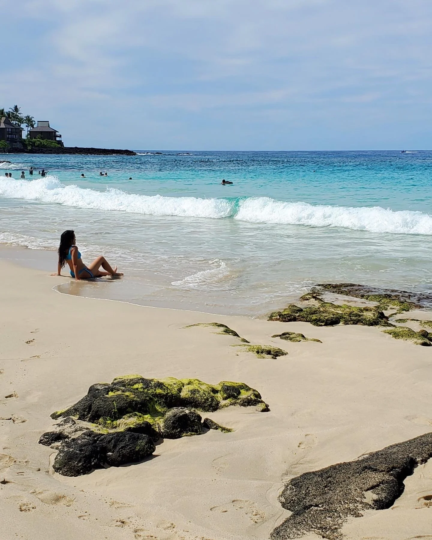 A woman in a blue swimsuit sitting on the sandy beach near rocks, with ocean waves and several people swimming in the background.