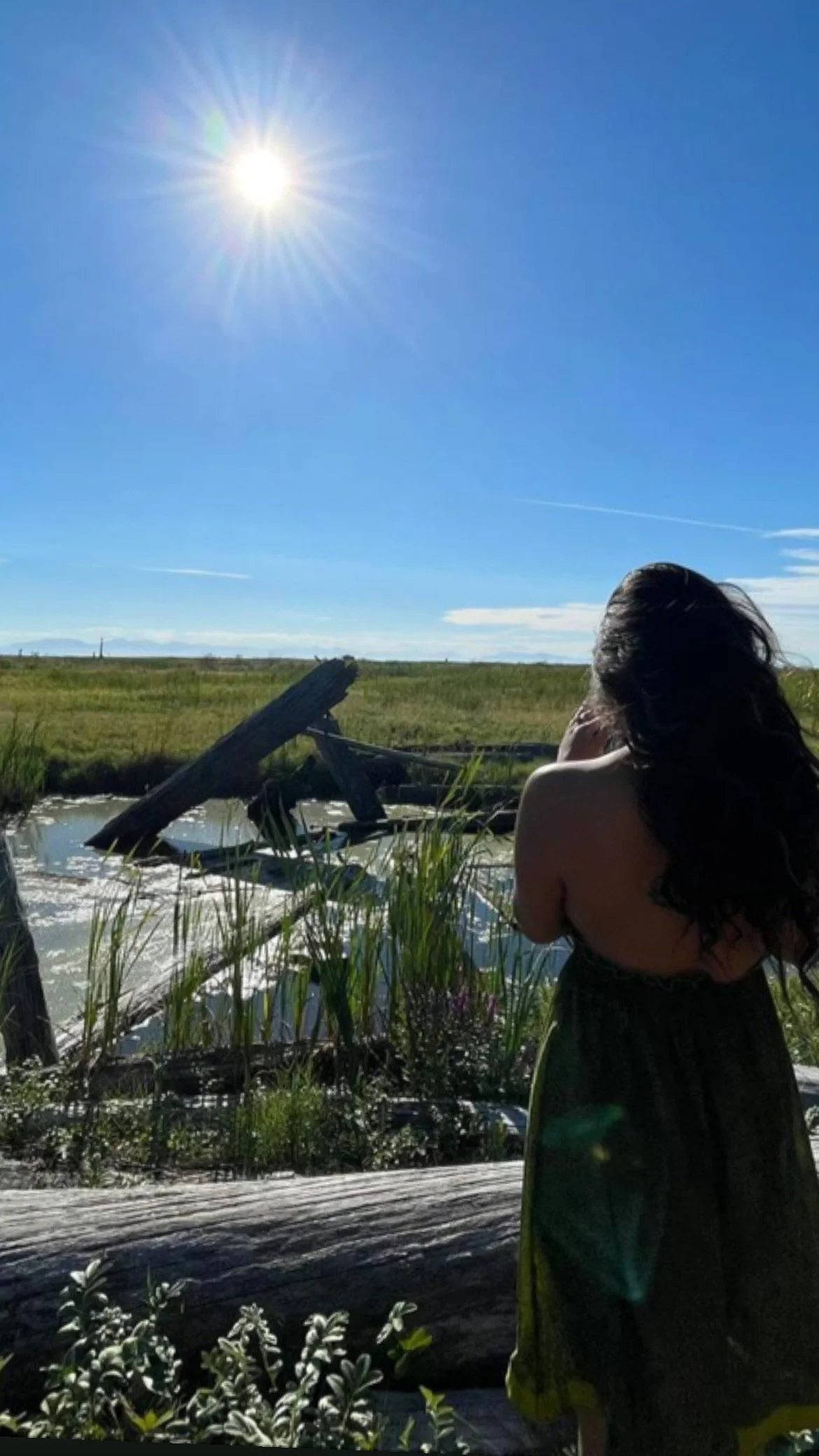 A woman with long dark hair standing near a waterway on a sunny day, with a partly broken wooden structure in the water and a clear blue sky.