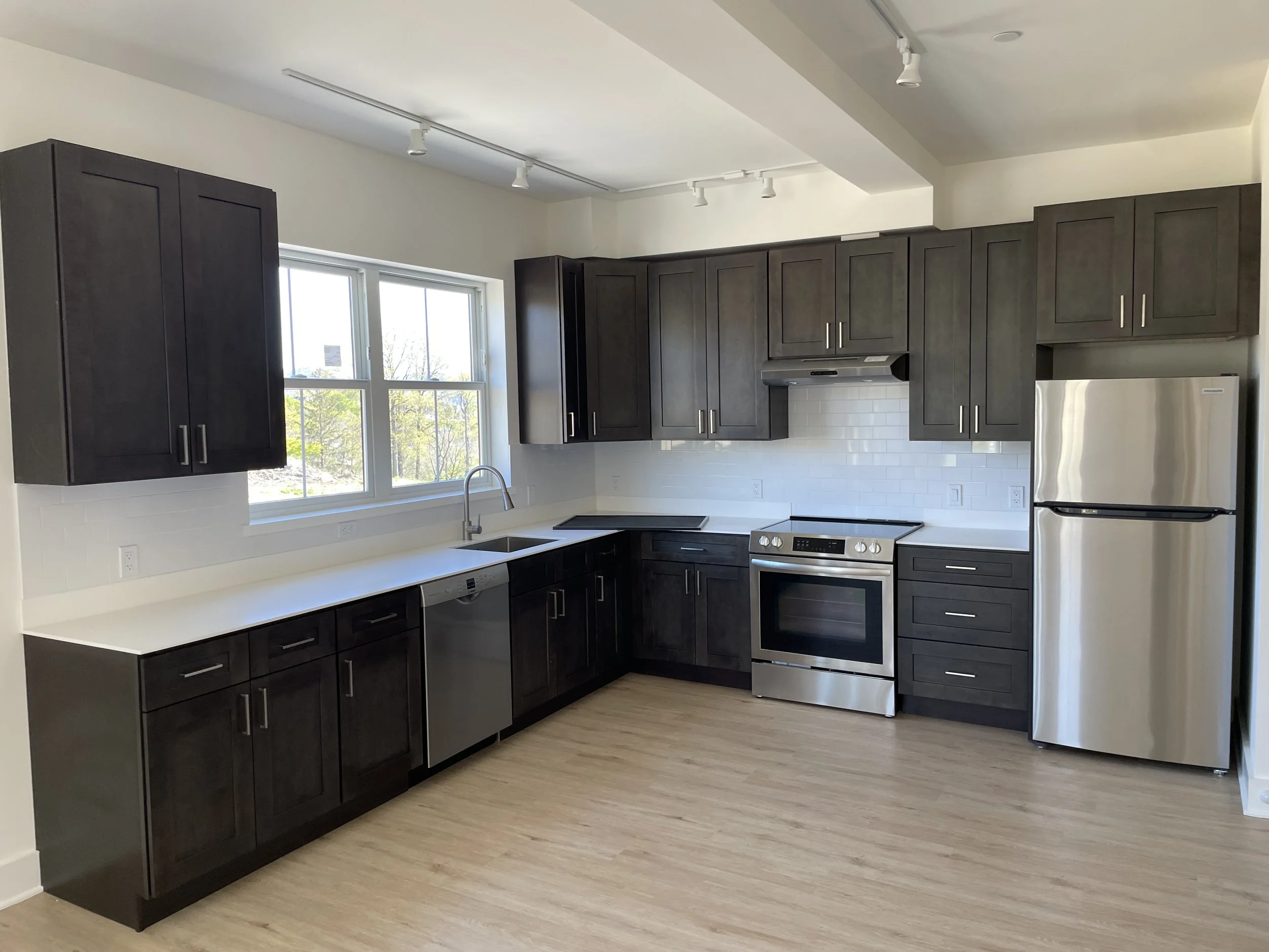 Modern kitchen with dark wood cabinets, white countertops, stainless steel appliances including a refrigerator and stove, white subway tile backsplash, and a window above the sink with a view of trees outside.