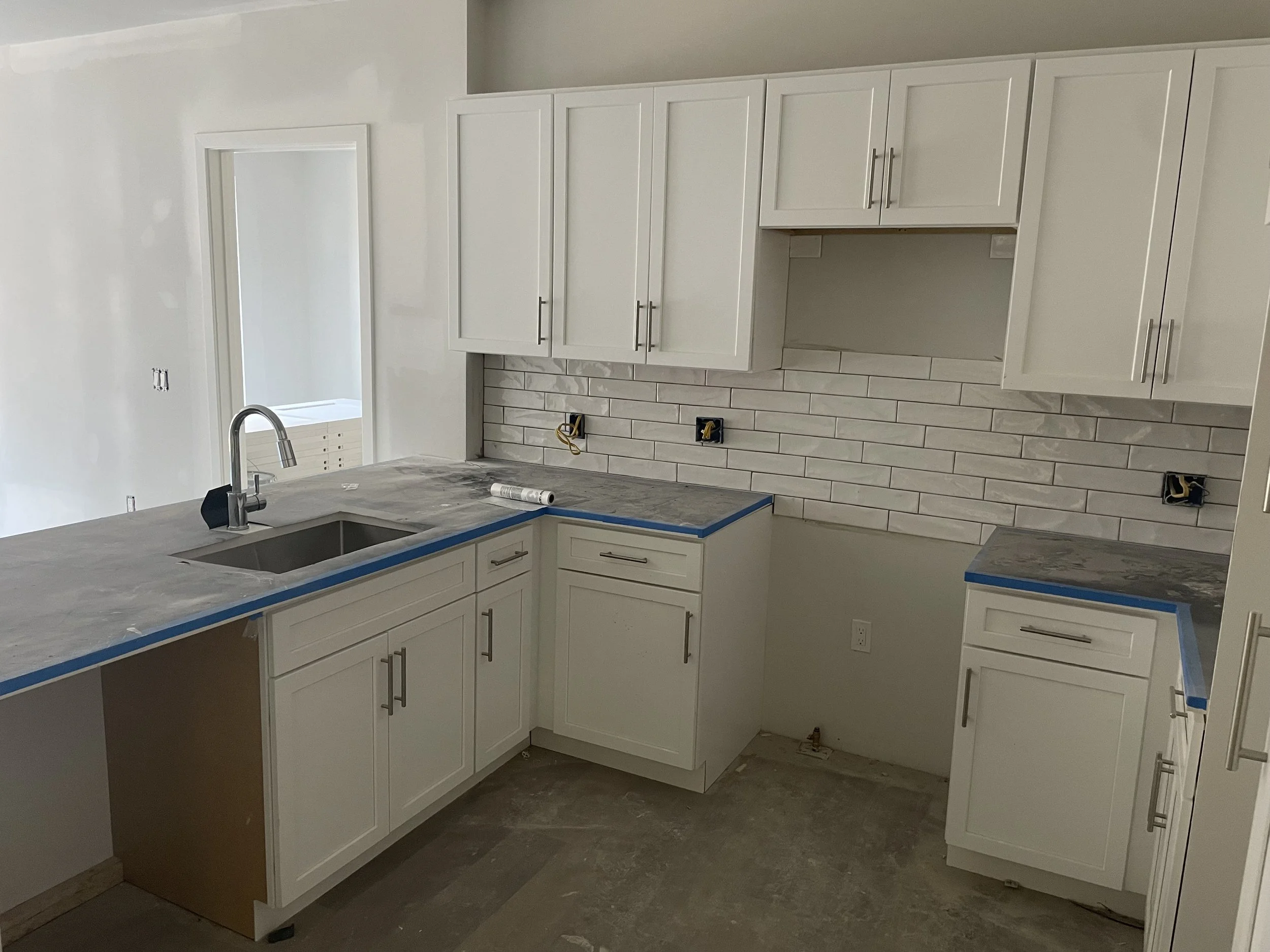 Kitchen under construction with white cabinets, a stainless steel sink, a marble countertop with blue painter's tape, and a brick-style tile backsplash.