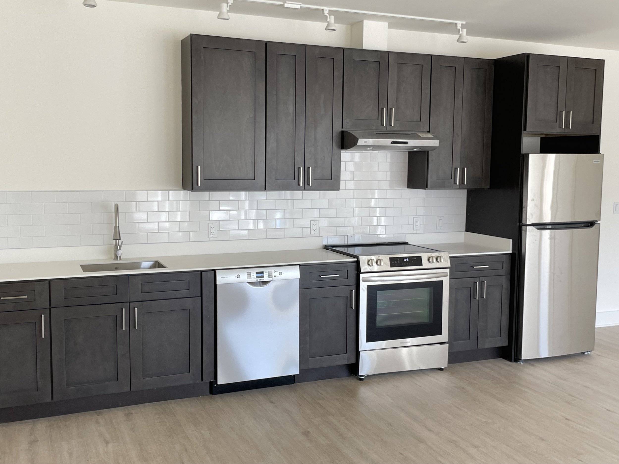 Modern kitchen with dark gray cabinets, stainless steel appliances including refrigerator, oven, and dishwasher, white subway tile backsplash, and a light-colored countertop.