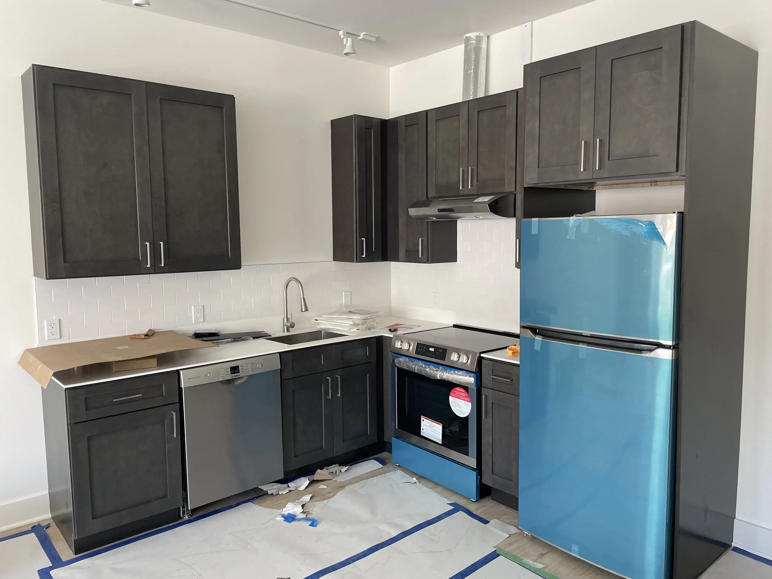 Kitchen with dark cabinets, stainless steel dishwasher, gas oven, and blue refrigerator, under construction or renovation with protective covering and construction debris on the floor.
