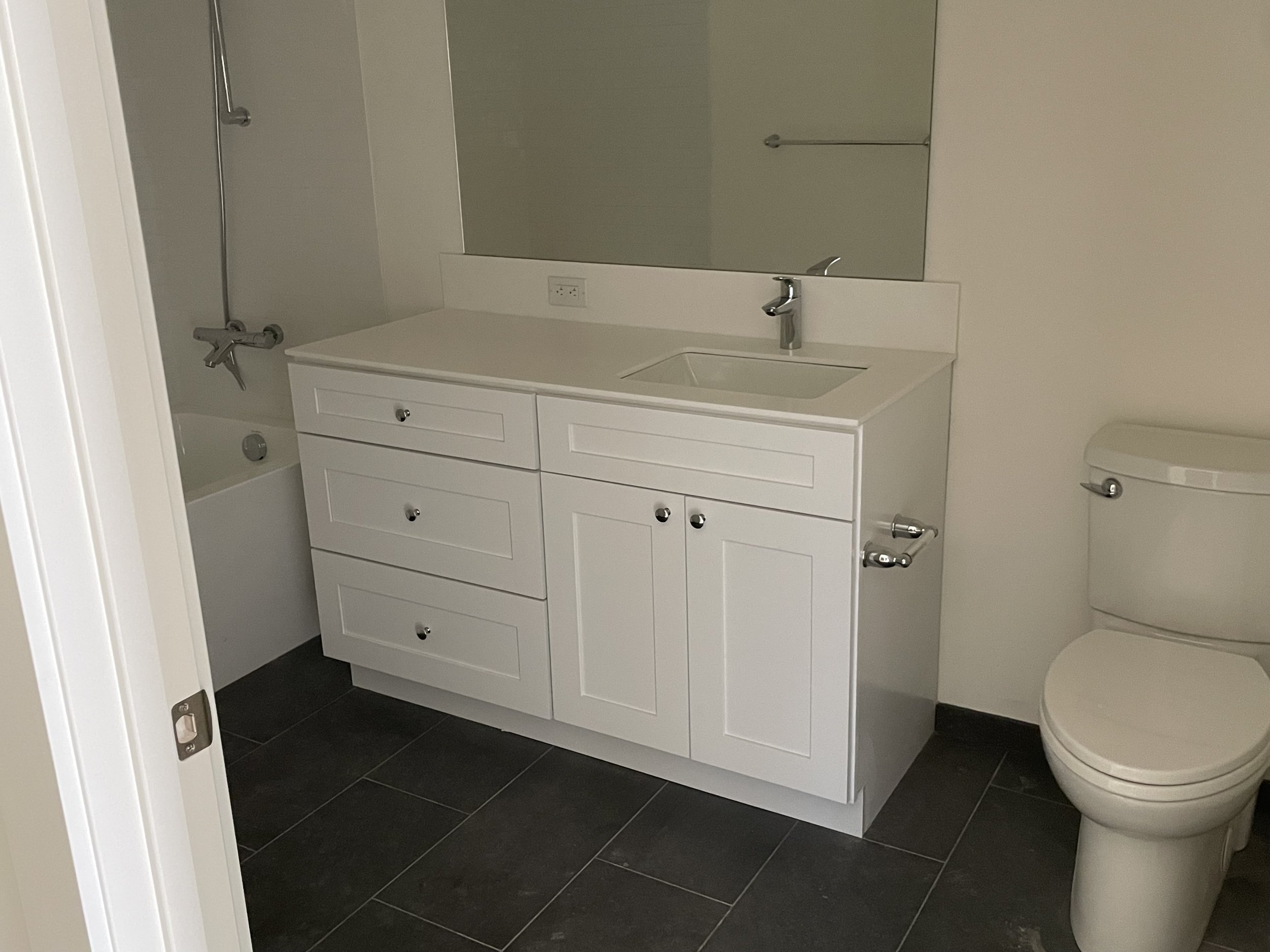 Bathroom with a white vanity, attached sink, and a mirror above. There is a bathtub with a shower and a toilet next to the vanity. The floor is tiled with dark tiles.