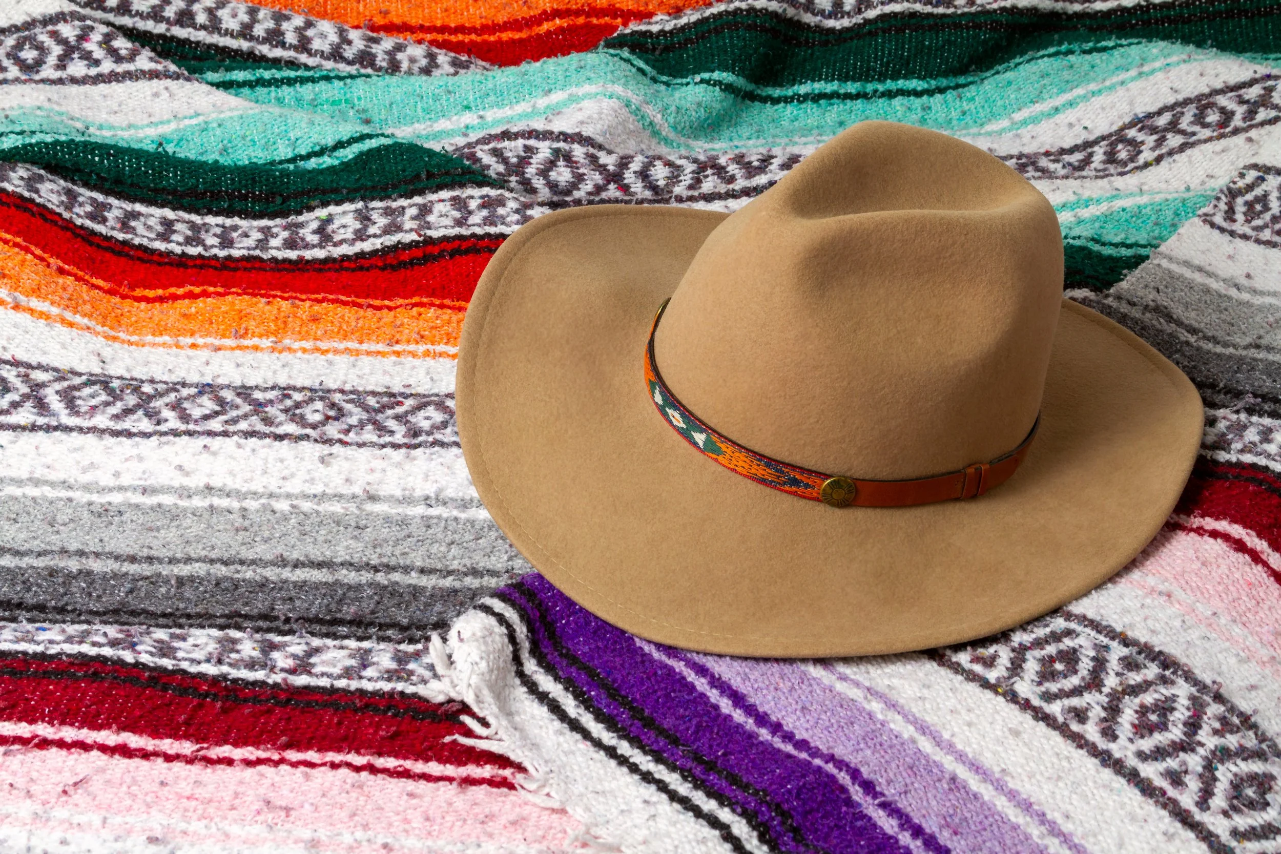A tan cowboy hat with a colorful band resting on a multicolored woven textile blanket.