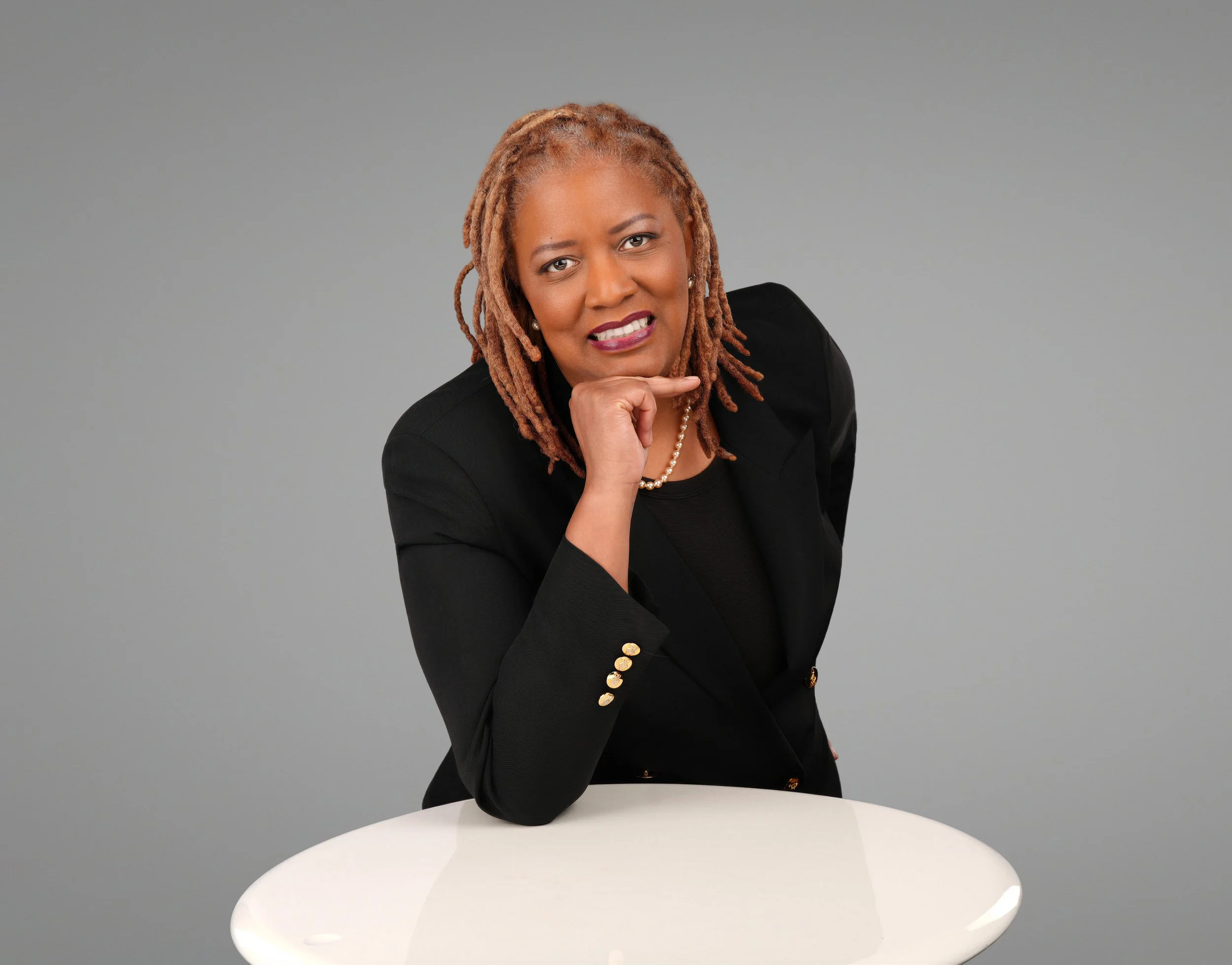 Yamila Williams  Psychiatric Nurse practitioner smiling, wearing shoulder-length  hair wearing a black blazer sitting at a white round table, resting her chin on her right hand, in front of a plain gray background.