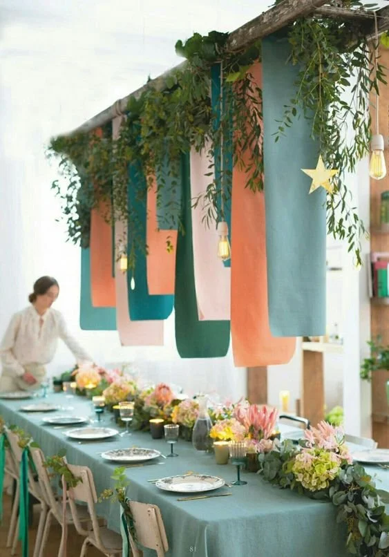 Elegant dining table setup with green, orange, and teal hanging fabric decorations, greenery, and floral centerpieces.