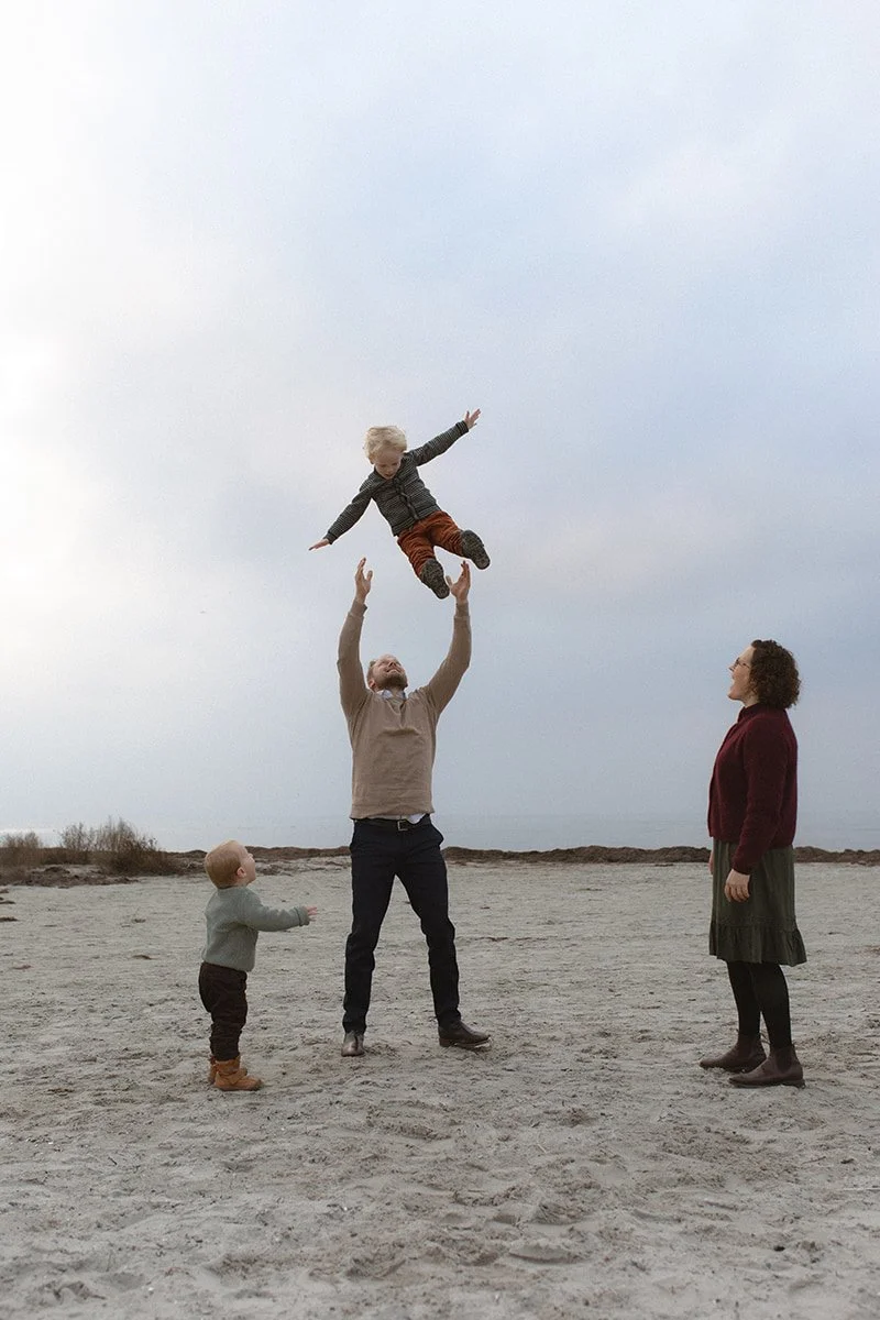 Pappa lyfter sitt barn i luften på stranden i Lomma under en mycket mulen eftermiddag.