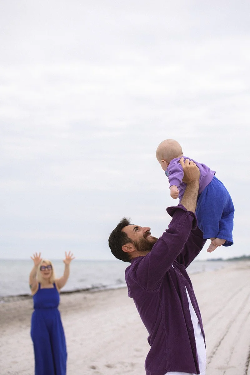 Pappa lyfter sin bebis i luften på stranden i Falsterbo under en ljus sommarmorgon i juni.
