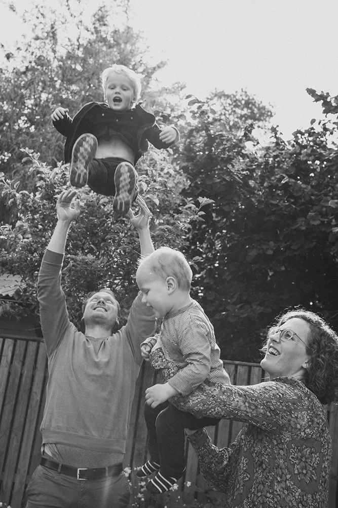 Familjefotografering i trädgården där pappa lyfter barnen i luften, naturliga och lekfulla familjebilder i Skåne