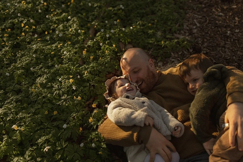 Pappa ligger i gräset med två barn på blommande äng under familjefotografering