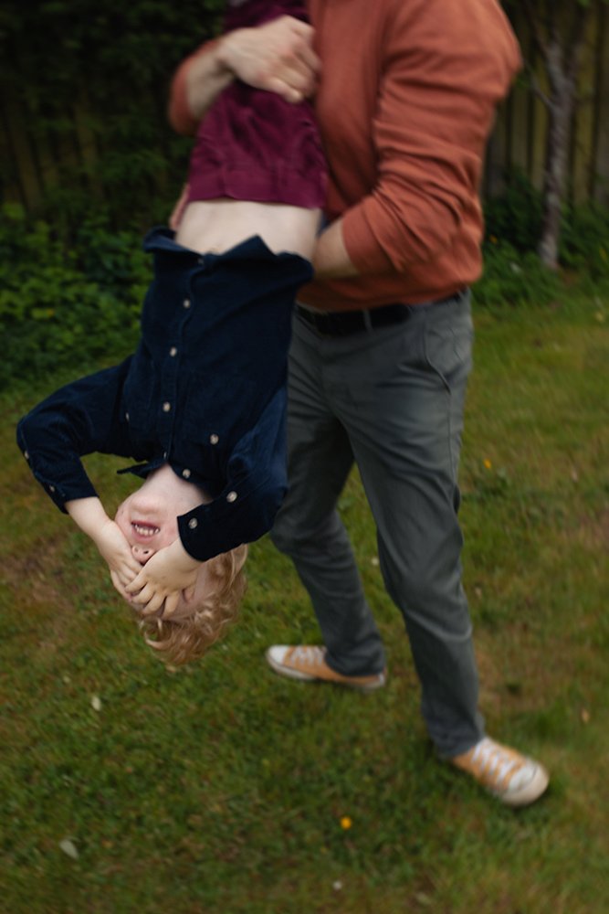 Naturlig familjefotografering där pappa leker med barn i trädgården, vardagliga ögonblick fångade utomhus i Skåne