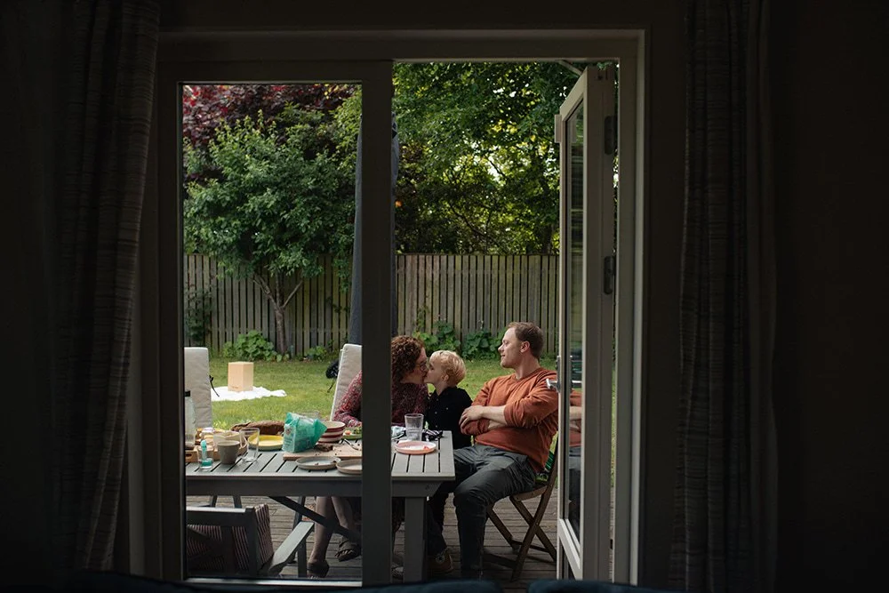 Familj samlad runt trädgårdsbordet sedd från insidan av huset, lifestyle familjefotografering hemma i Lund