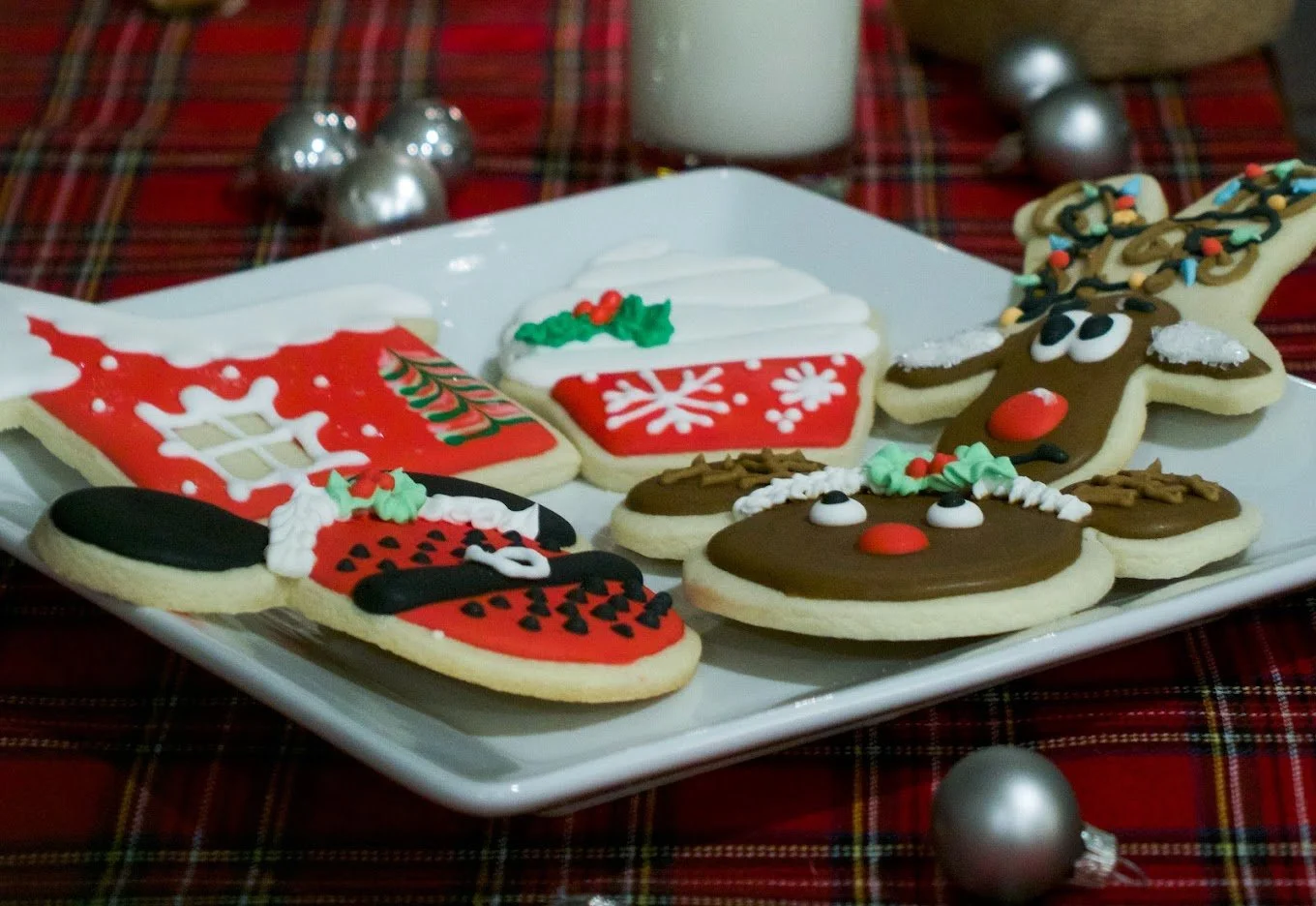Assorted Christmas-themed decorated sugar cookies on a white plate, including reindeer, Santa hat, Christmas tree, mitten, and snowflake designs, with a red and green plaid tablecloth in the background.
