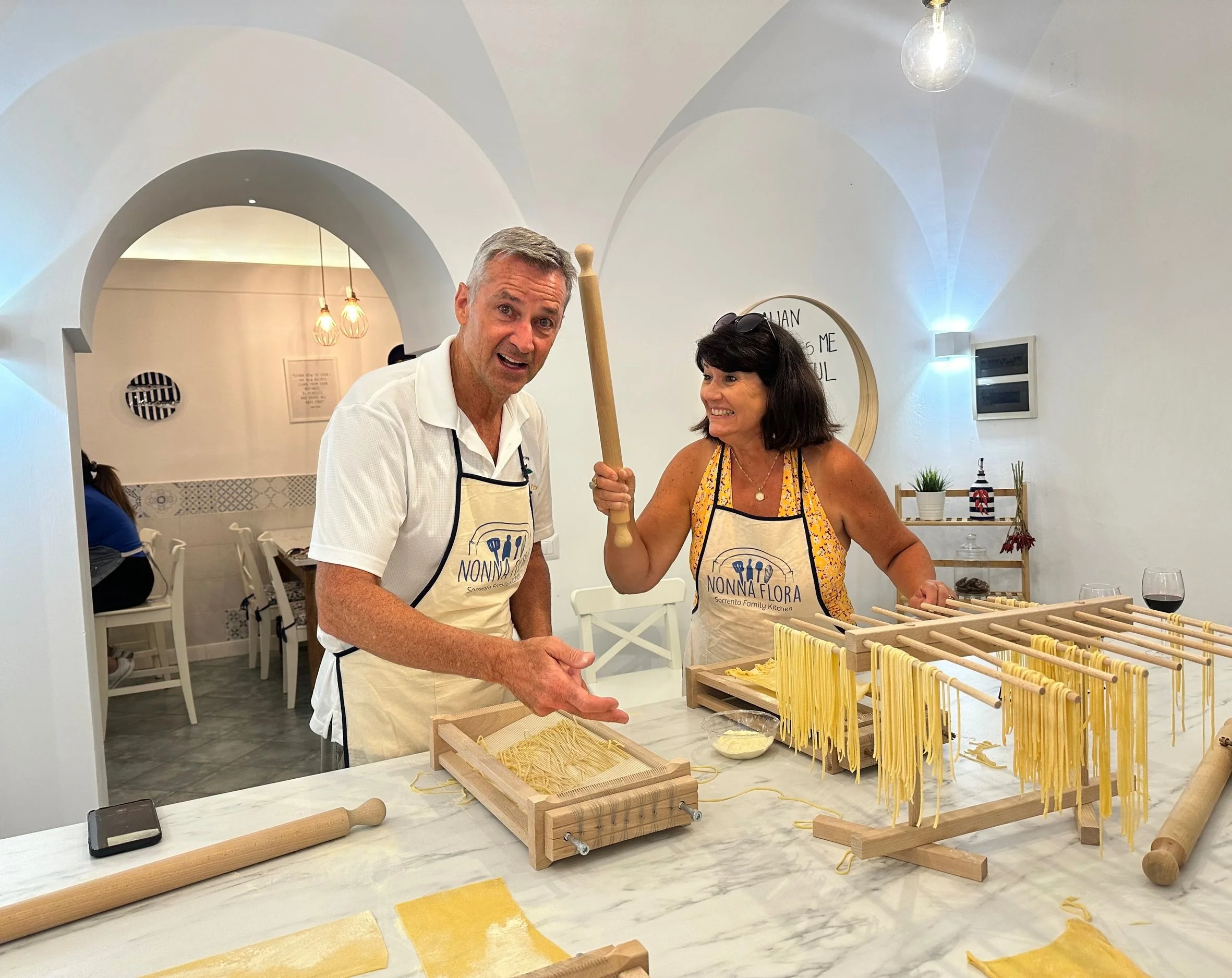 Two people with aprons baking in a bright kitchen. The woman holds a rolling pin and the man points at baked goods on table.