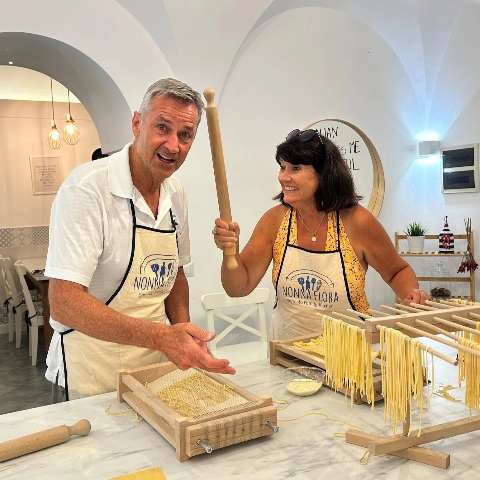 Two people with aprons baking in a bright kitchen. The woman holds a rolling pin and the man points at baked goods on table.