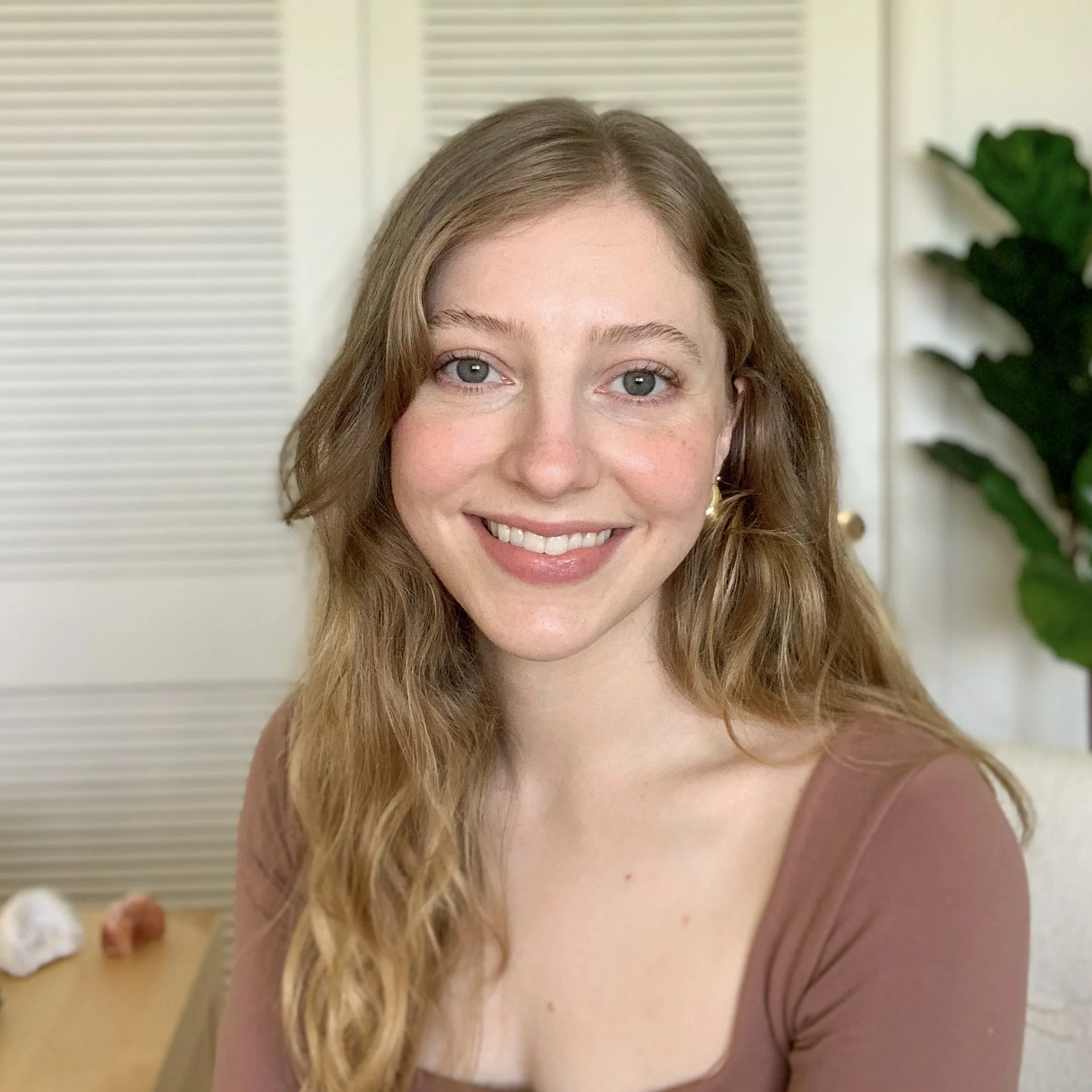 A young woman with long, wavy, light brown hair and blue eyes, smiling indoors with a white door and green plant in the background.