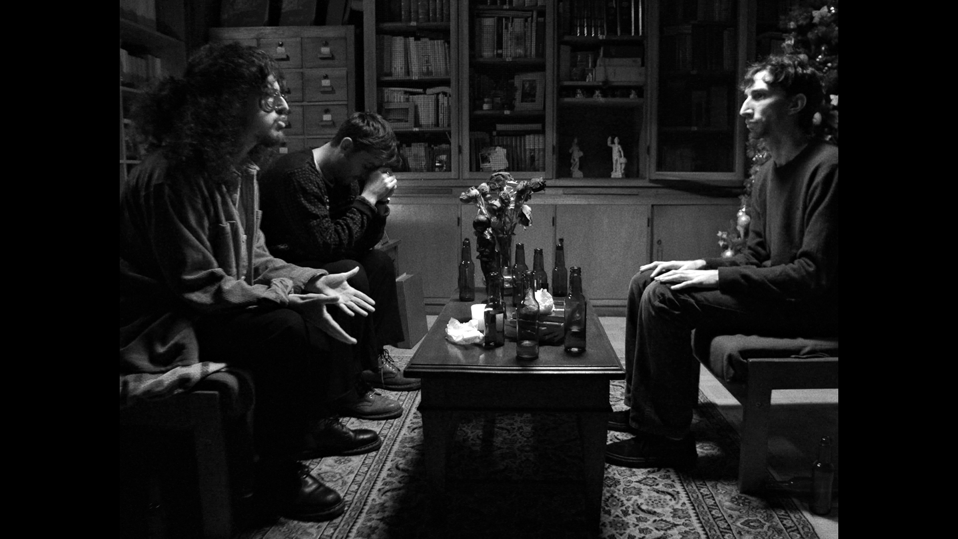 Quatro pessoas sentadas em volta de uma mesa de centro, em uma sala com móveis de madeira e livros na estante ao fundo, numa cena em preto e branco. Dois homens na esquerda parecem estar discutindo ou explicando algo, enquanto o homem à direita escuta atentamente. Há garrafas de vidro na mesa.