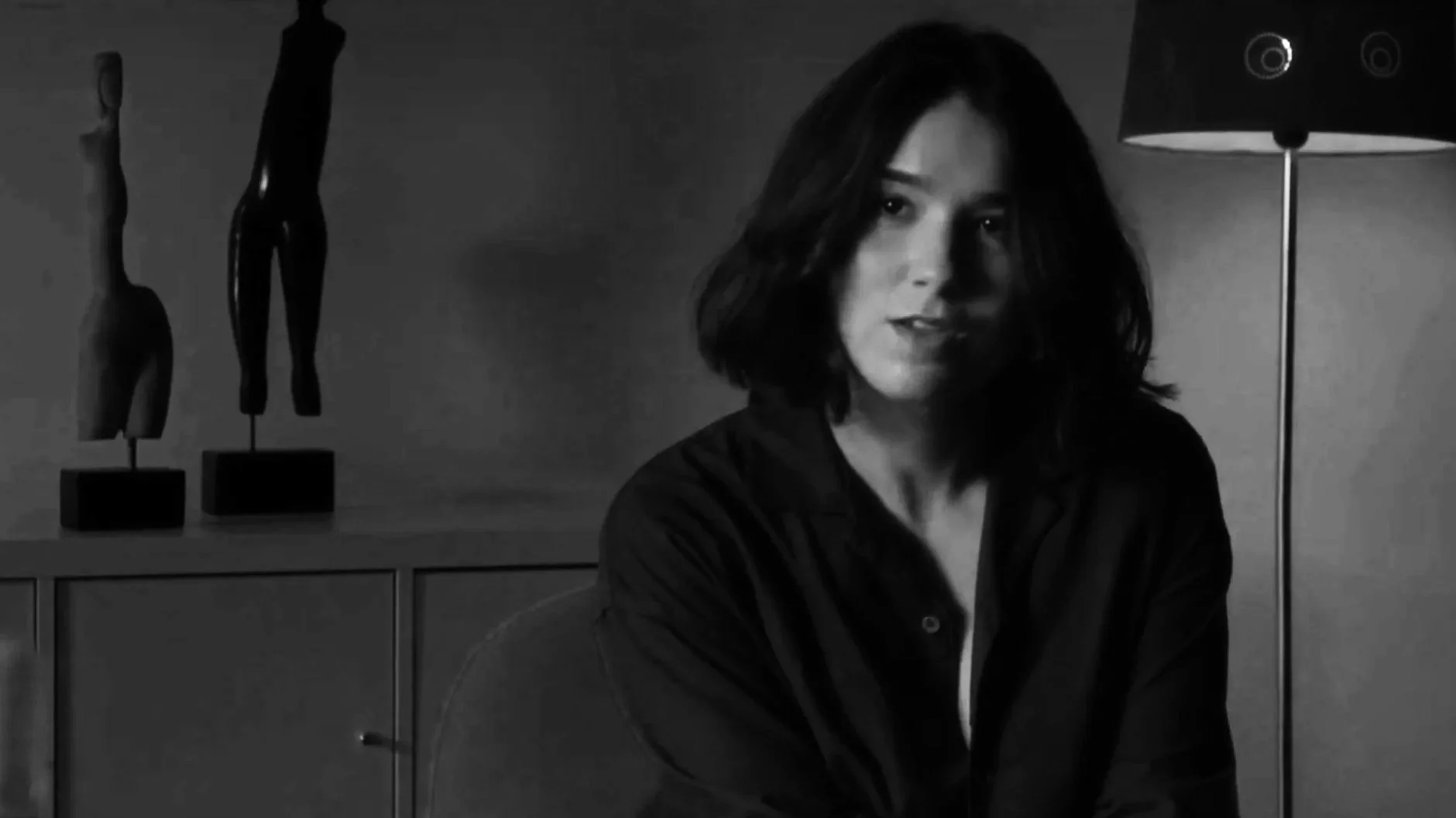 Retrato de uma mulher de cabelos castanhos escuros e cabelo na altura dos ombros, usando uma blusa escura, em um ambiente com uma lâmpada de pé ao lado e estátua de uma figura feminina na parede ao fundo, em preto e branco.