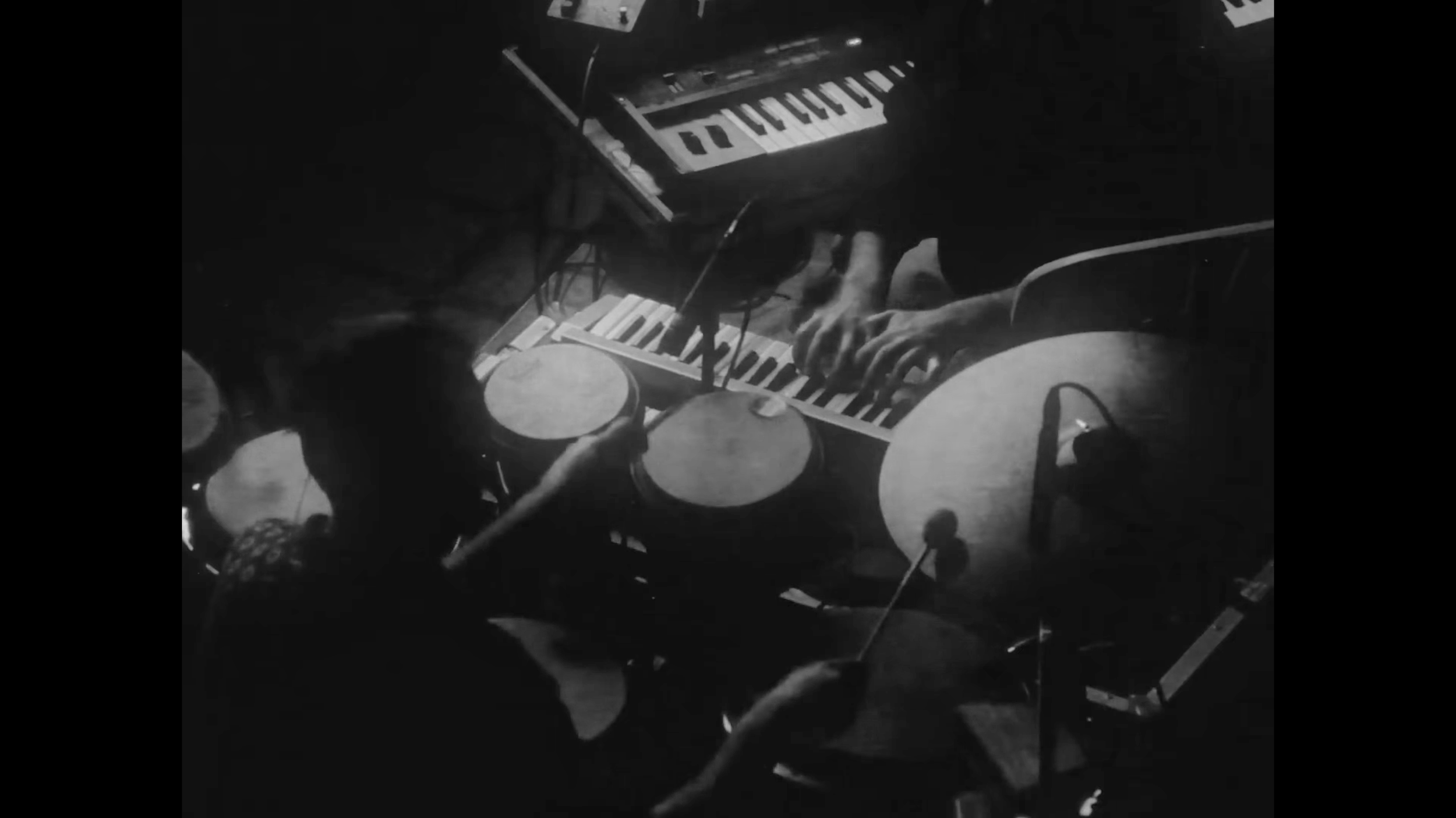 Pessoas tocando instrumentos musicais em um palco, incluindo um teclado, um piano e instrumentos de percussão, em uma apresentação ao vivo