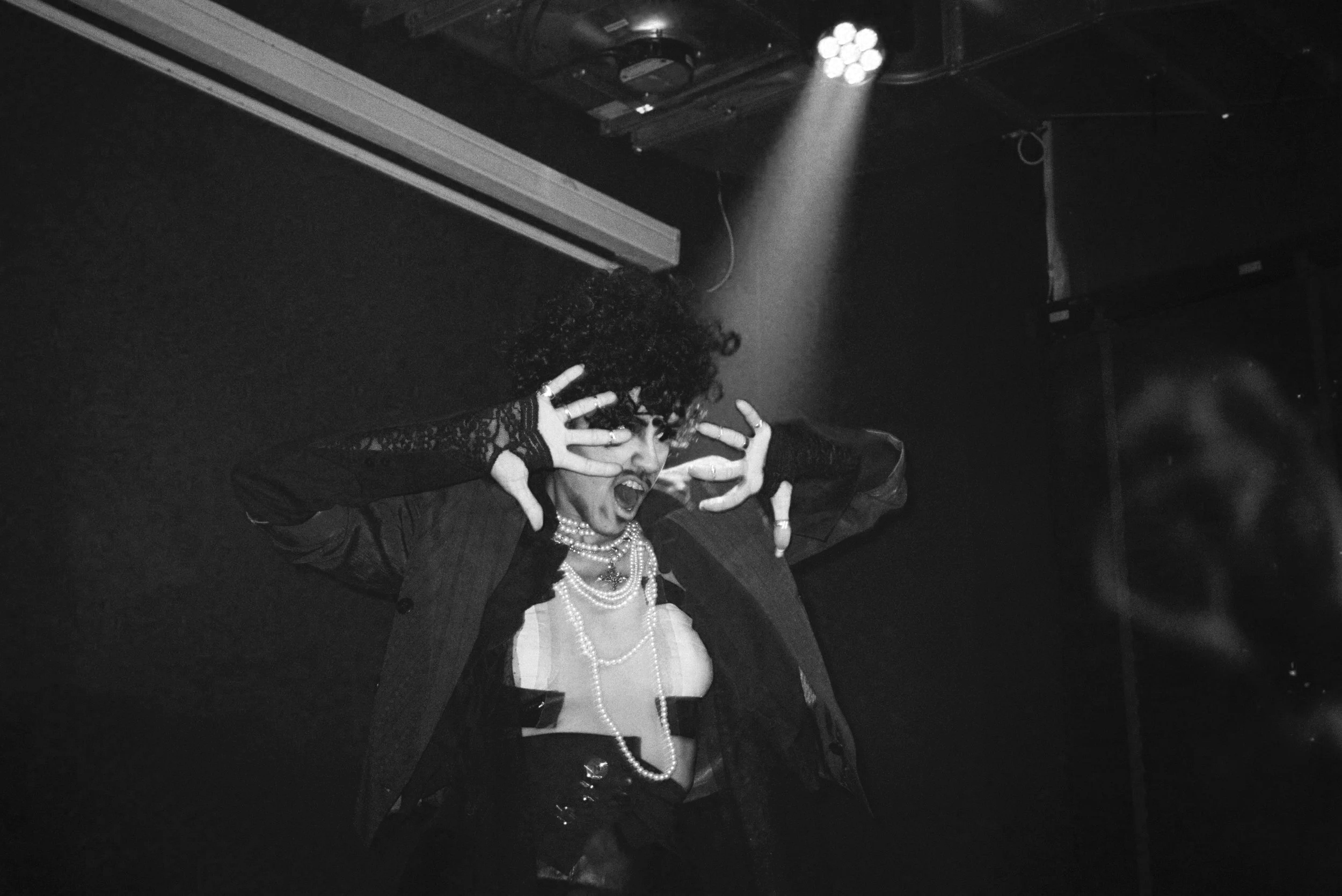 Pessoa performando no palco, com roupas extravagantes, brincos de pérolas, making facial expressões dramáticas, iluminação de palco.